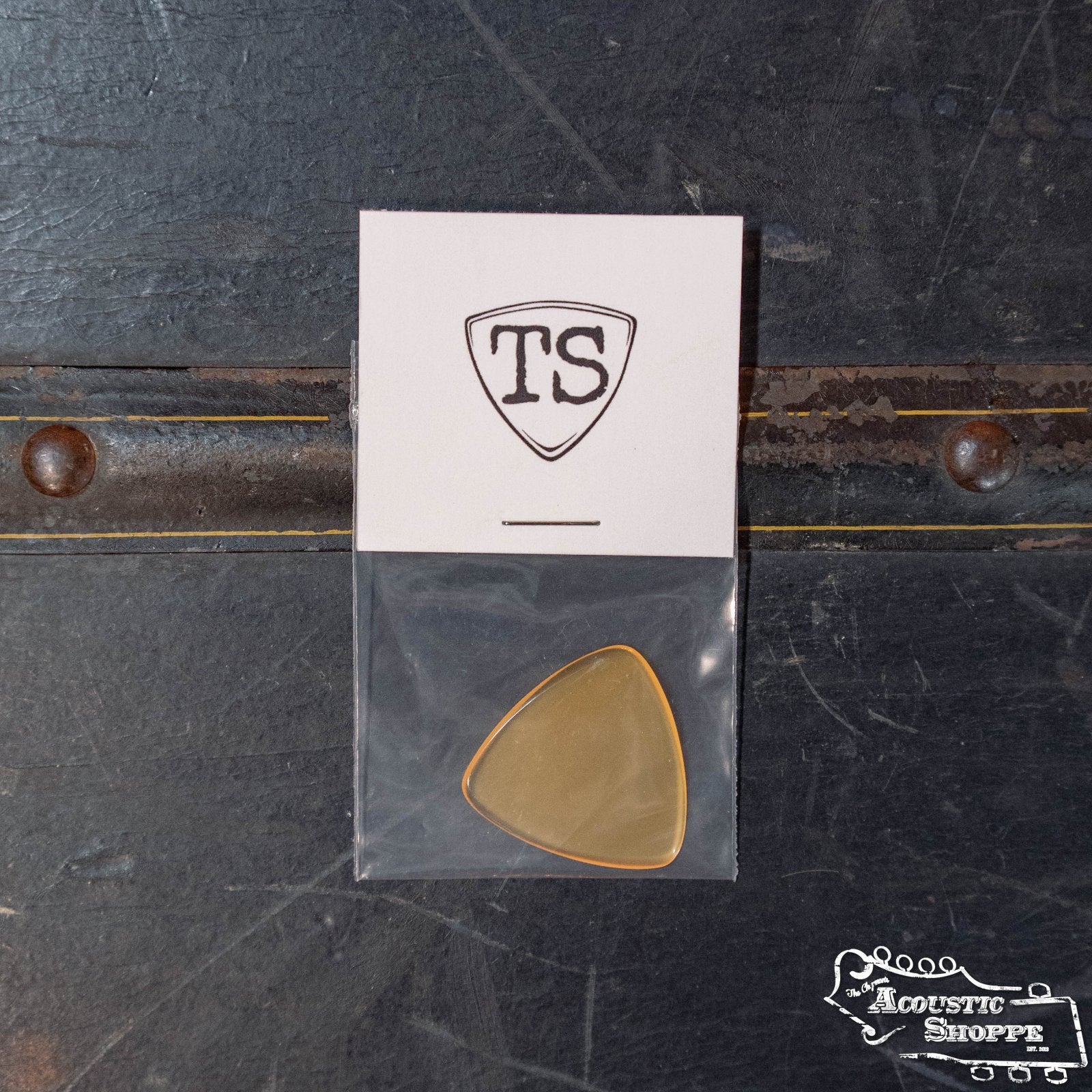 A ToneSlabs Tweener Triangle Guitar Pick - 1.5 mm by ToneSlabs, in a clear pouch on a white “TS” logo card, rests on a dark textured surface, with The Acoustic Shoppe logo in the bottom right corner.