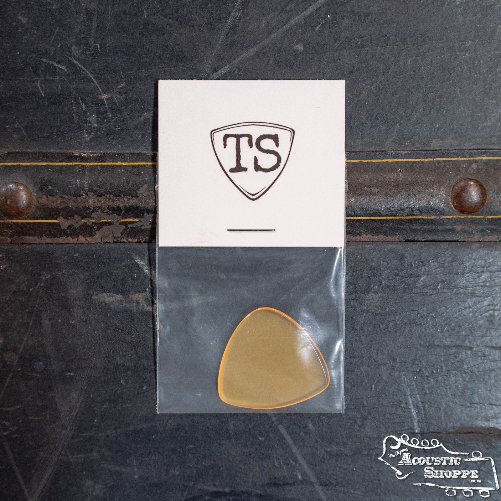 A ToneSlabs Tweener Triangle w/One Rounded Edge Guitar Pick (1.5 mm) in a plastic bag sits on a dark surface. A white card with the TS shield logo is above it, and “The Acoustic Shoppe” logo appears at the lower right.