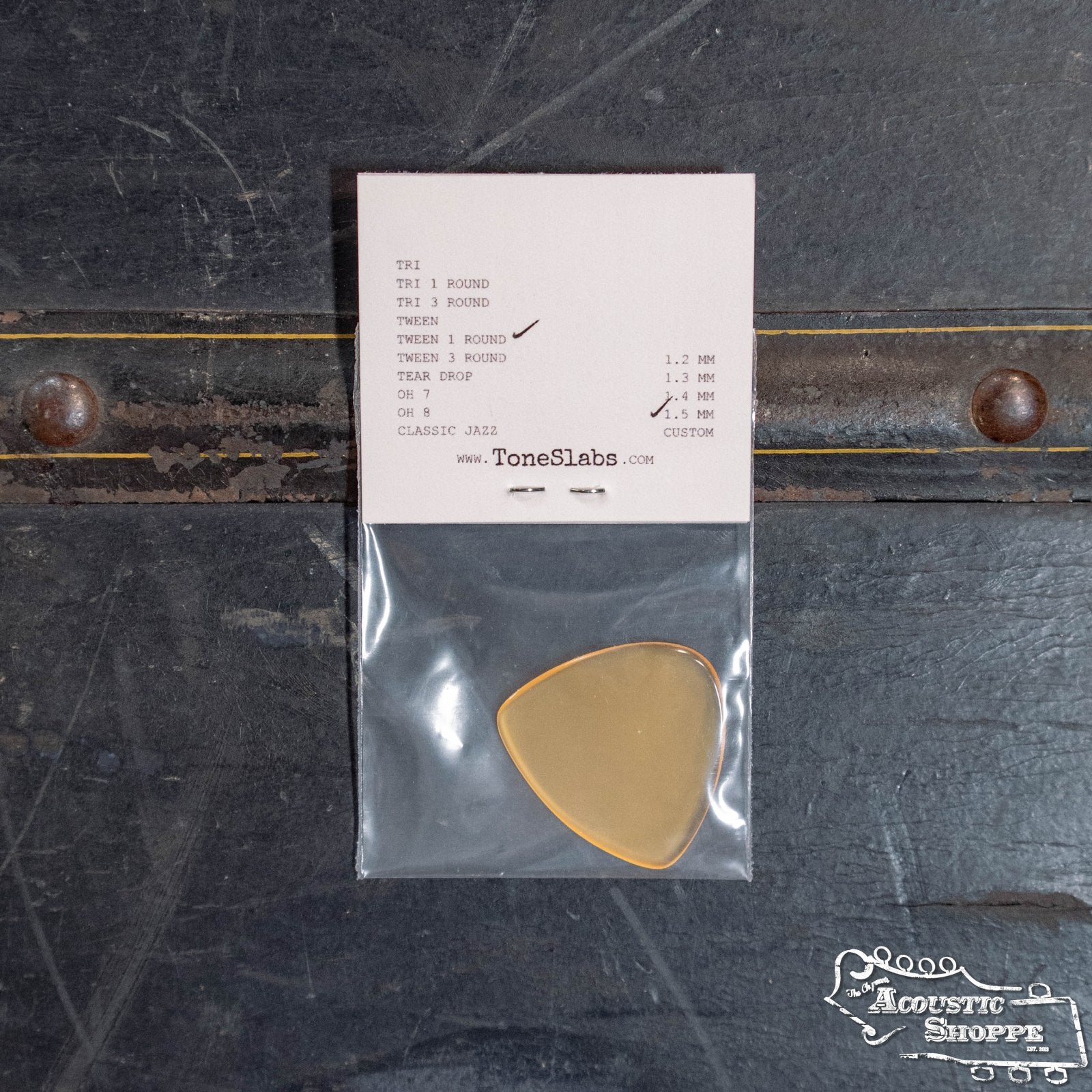 A translucent ToneSlabs Tweener Triangle w/One Rounded Edge Guitar Pick (1.5 mm) is shown in a clear plastic bag on a dark surface, with a white ToneSlabs card detailing size, thickness, and brand above the pick.