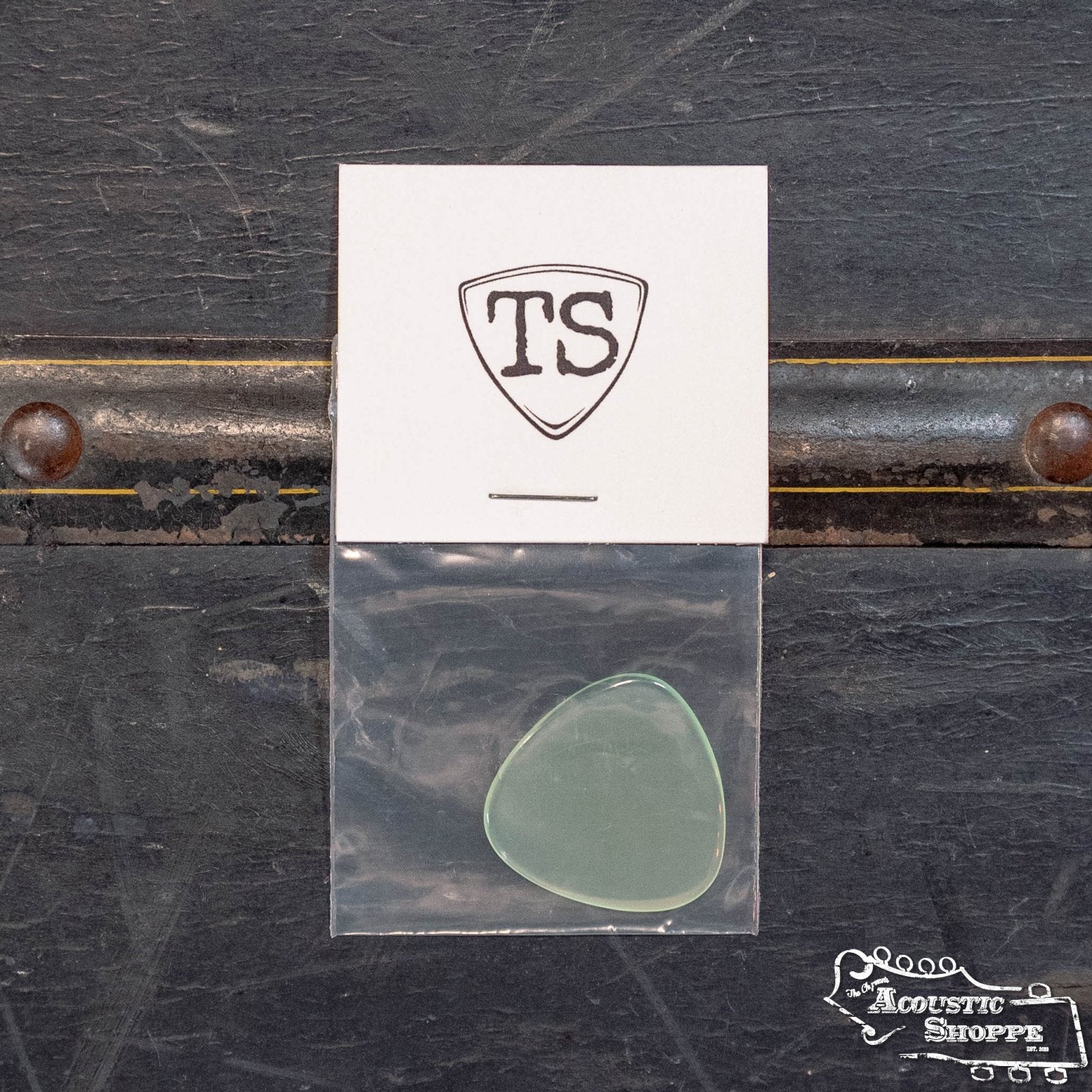 A translucent green ToneSlabs Tweener Triangle 3Rounded Guitar Pick (1.3 mm) is packaged in a plastic bag stapled to a white card with a TS shield. The card sits on black, displaying The Acoustic Shoppe logo at the bottom right.