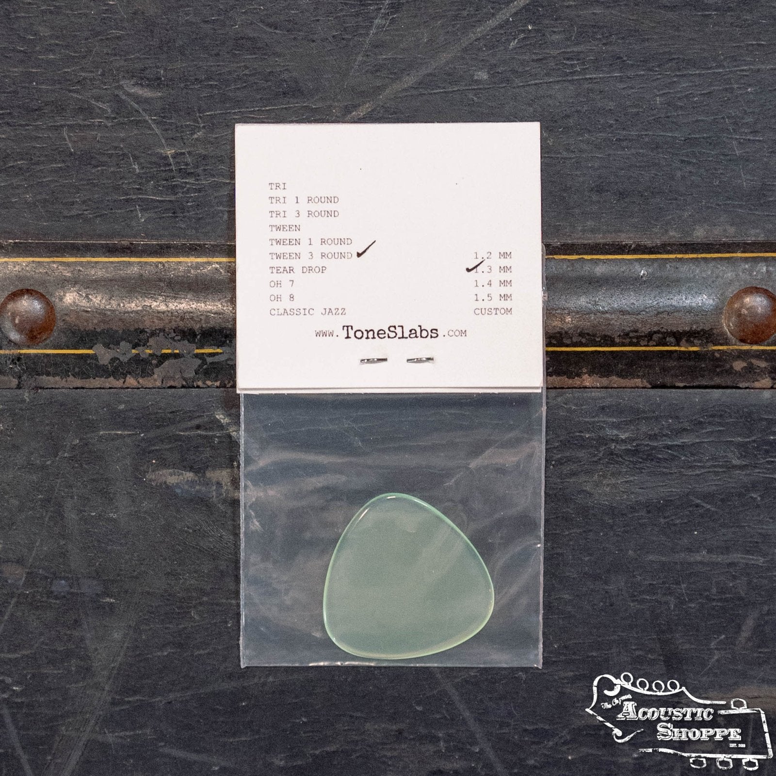 A translucent ToneSlabs Tweener Triangle 3Rounded Guitar Pick (1.3 mm) in a plastic bag lies beneath its spec card on a dark textured surface featuring The Acoustic Shoppe logo at the lower right.