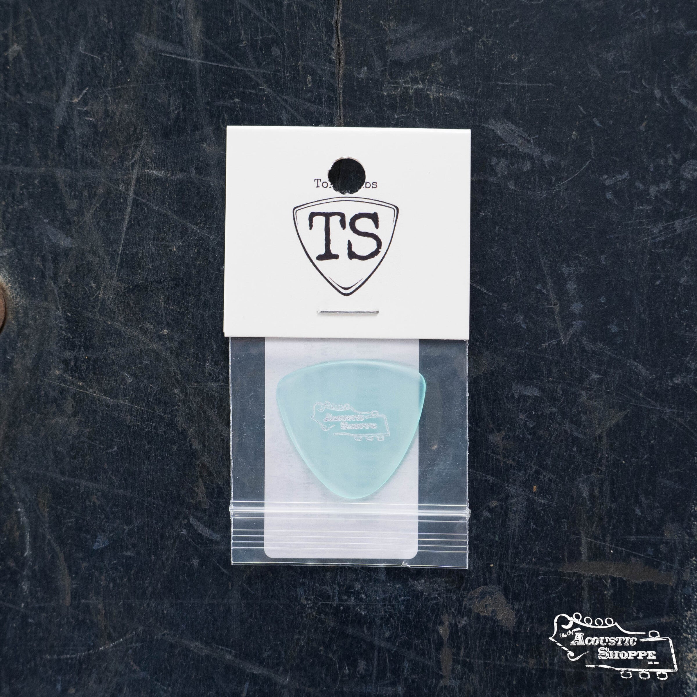 A ToneSlabs Tweener Triangle w/ One Rounded Edge Guitar Pick (1.3mm, light blue) is in a small clear bag on a white card with the TS shield logo, set on a dark textured surface. “Road Acoustic Shoppe” appears at the bottom right.
