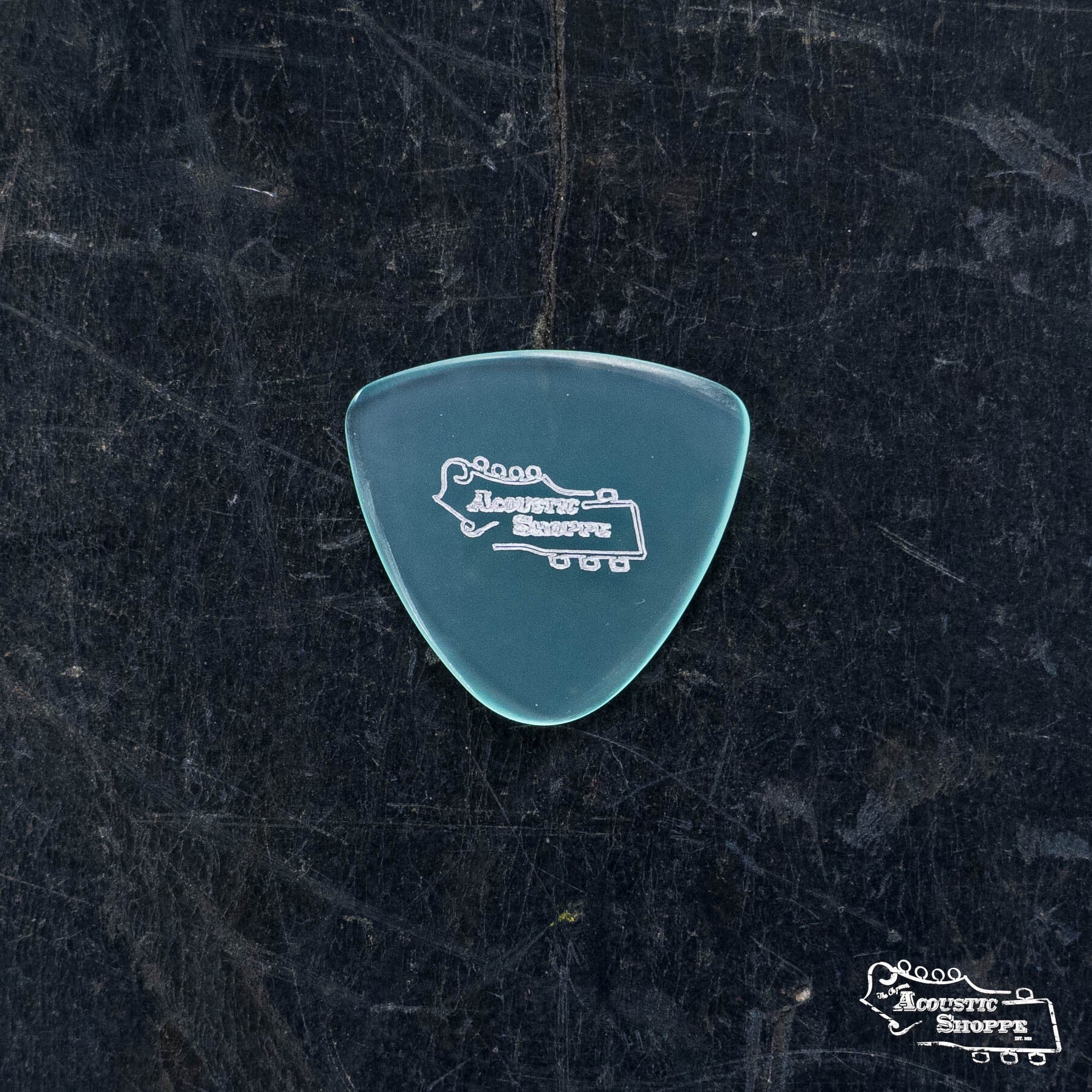A translucent turquoise ToneSlabs Tweener Triangle w/One Rounded Edge Guitar Pick (1.3mm) with a white Acoustic Shoppe logo rests on a dark textured surface, with a smaller logo in the lower right corner.