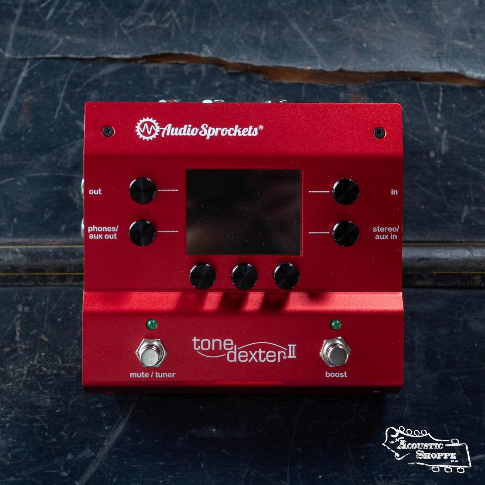 A red Audio Sprockets ToneDexter II acoustic preamp pedal with black knobs, a digital screen, and two footswitches rests on a dark, scratched surface. The Acoustic Shoppe logo is visible in the lower right corner.