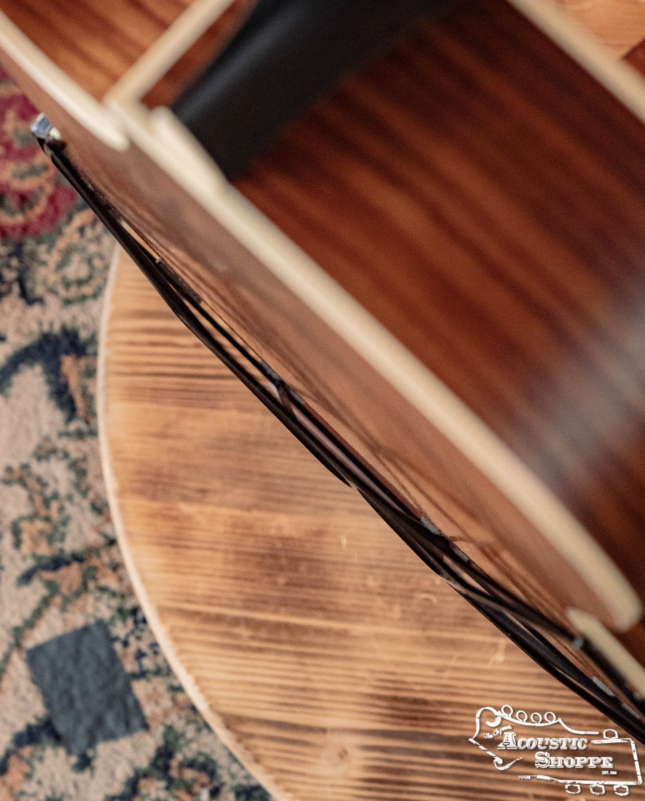 Close-up of a guitars side and edge on wood, patterned carpet in view. The Acoustic Shoppe logo appears at lower right. The Tone-Gard Dreadnaught Guitar D-Gard by Tone-Gard can help enhance your instruments tone.