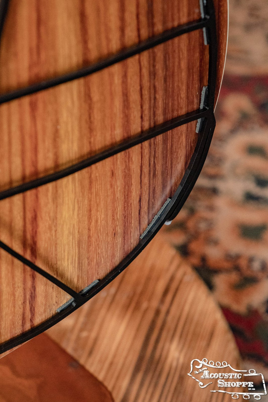 Close-up of a wooden guitar body with a black metal Tone-Gard Dreadnaught Guitar D-Gard frame by Tone-Gard, set on a patterned rug. The Acoustic Shoppe logo in the corner highlights this accessorys ability to enhance your instrument’s tone.