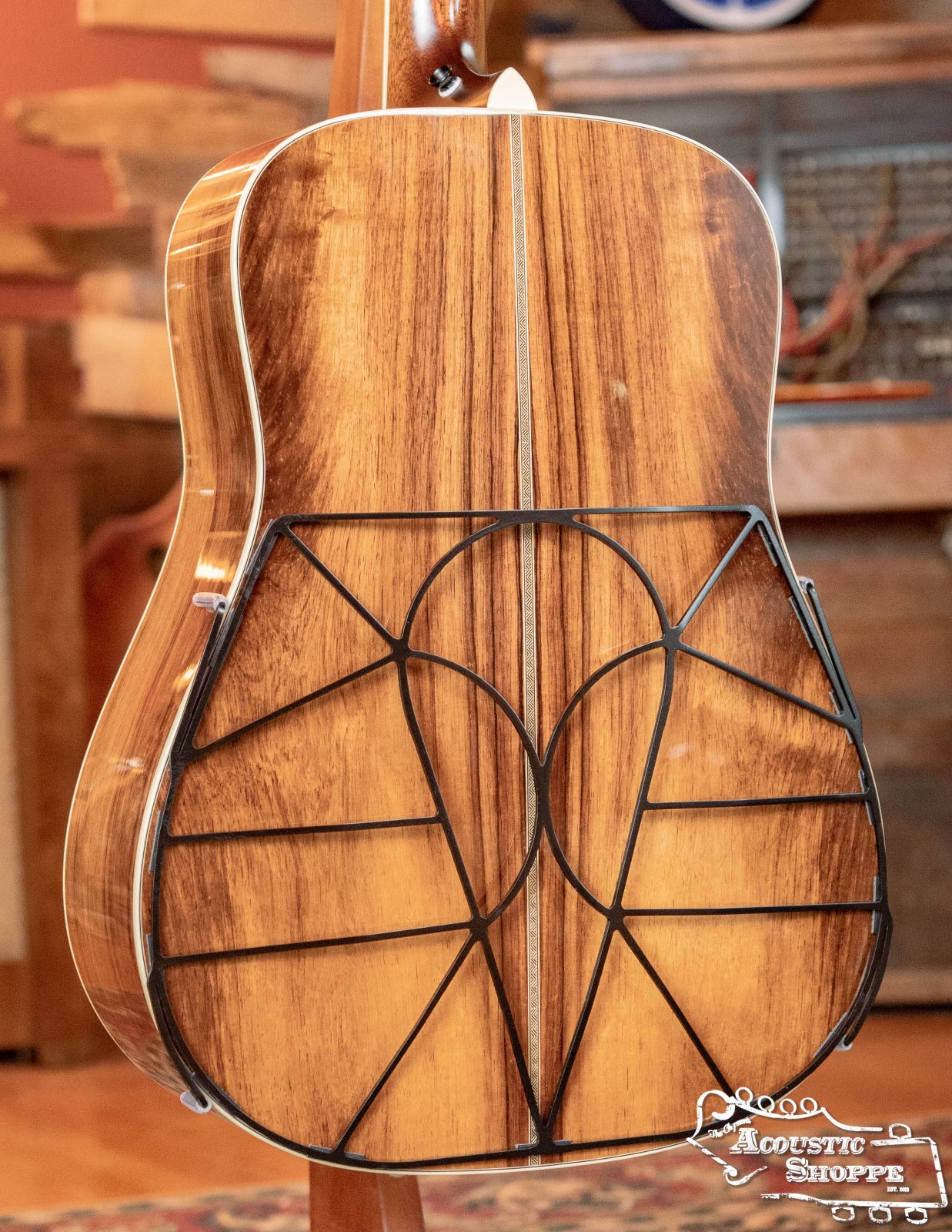 A Tone-Gard Dreadnaught Guitar D-Gard by Tone-Gard is shown fitted to the back of an acoustic guitar with a wood finish. Its unique geometric black metal frame enhances tone. The Acoustic Shoppe logo appears in the bottom right corner.