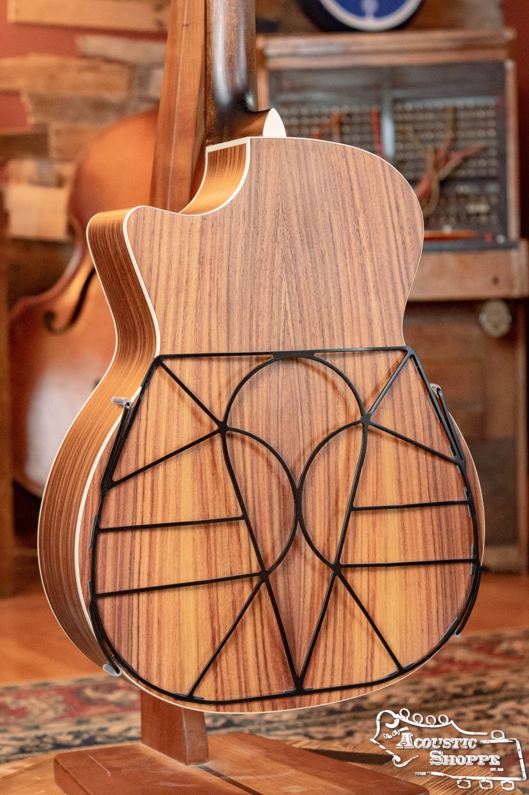 A wooden acoustic guitar with the Tone-Gard Dreadnaught Guitar D-Gard—a geometric black metal back accessory by Tone-Gard—stands upright in a cozy, rustic room with musical instruments, a patterned rug, and the Acoustic Shoppe logo in the corner.