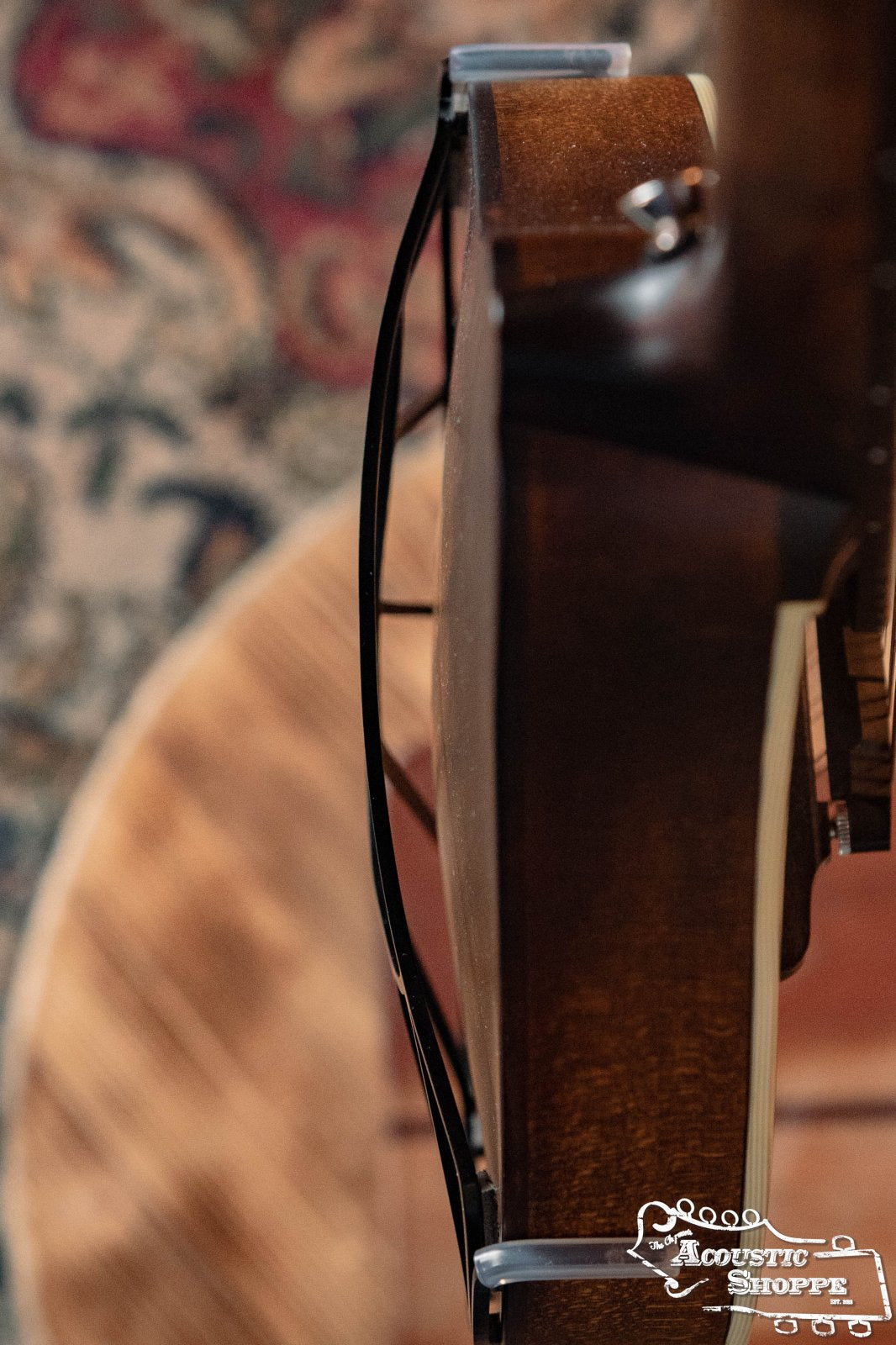 A close-up side view of an acoustic guitar with a visible crack along the body, set against a blurred patterned rug and wooden floor. The Acoustic Shoppe logo appears as in images featuring the Tone-Gard Mandolin - Sunrise by Tone-Gard.