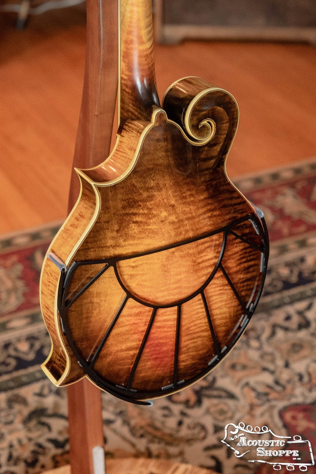 A close-up of a Tone-Gard Mandolin - Sunrise by Tone-Gard, featuring a glossy sunburst finish, detailed wood grain, ornate carving, and a back protector. It stands on a wooden stand above a patterned rug with The Acoustic Shoppe logo visible.