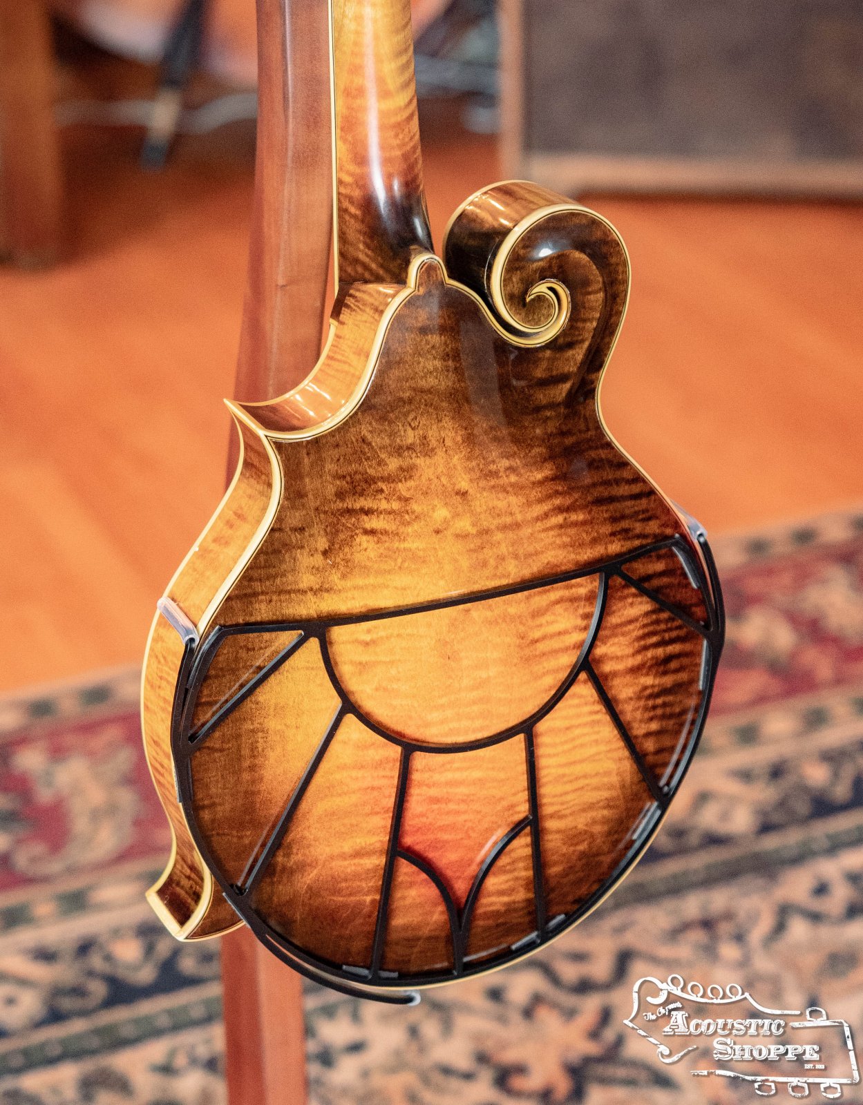 A close-up of the back of a mandolin with a sunburst finish and black line inlay showcases the Tone-Gard Mandolin - Vintage by Tone-Gard, standing on a patterned rug near an amplifier. The Acoustic Shoppe logo appears in the bottom right corner.