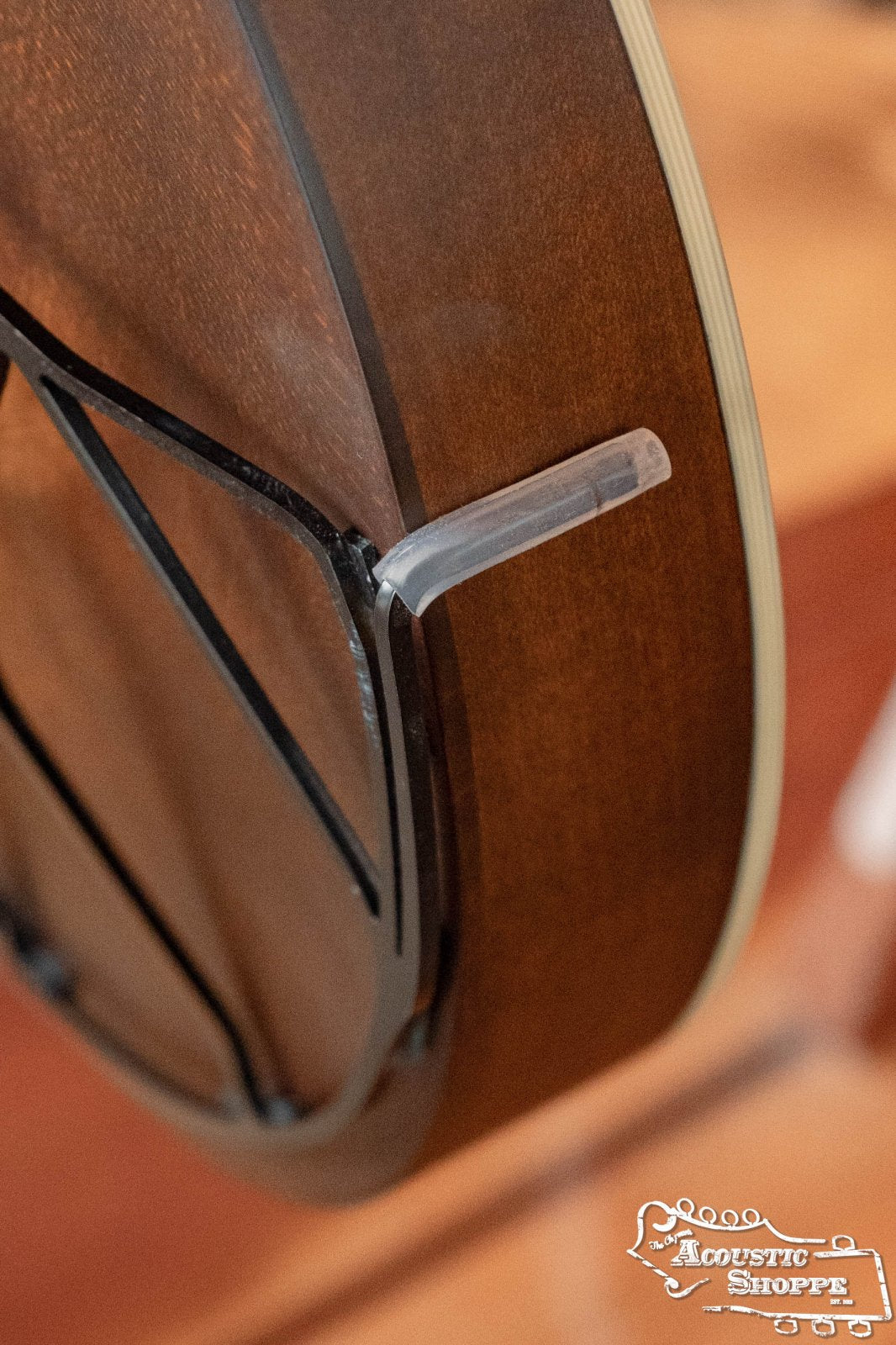 Close-up of a brown acoustic instrument fitted with a Tone-Gard OM-Gard Octave Mandolin tone enhancer by Tone-Gard, featuring a black metal armrest and transparent plastic tip, with Acoustic Shoppe branding in the lower right corner.