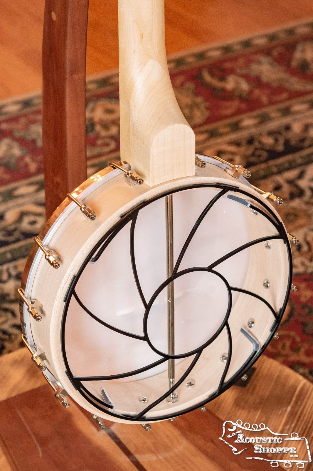 A Tone-Gard Open Back Banjo (12) by Tone-Gard with a light wood finish and black spiral metal design across the clear drumhead rests on a wooden stand. A patterned rug and The Acoustic Shoppe logo appear in the background.