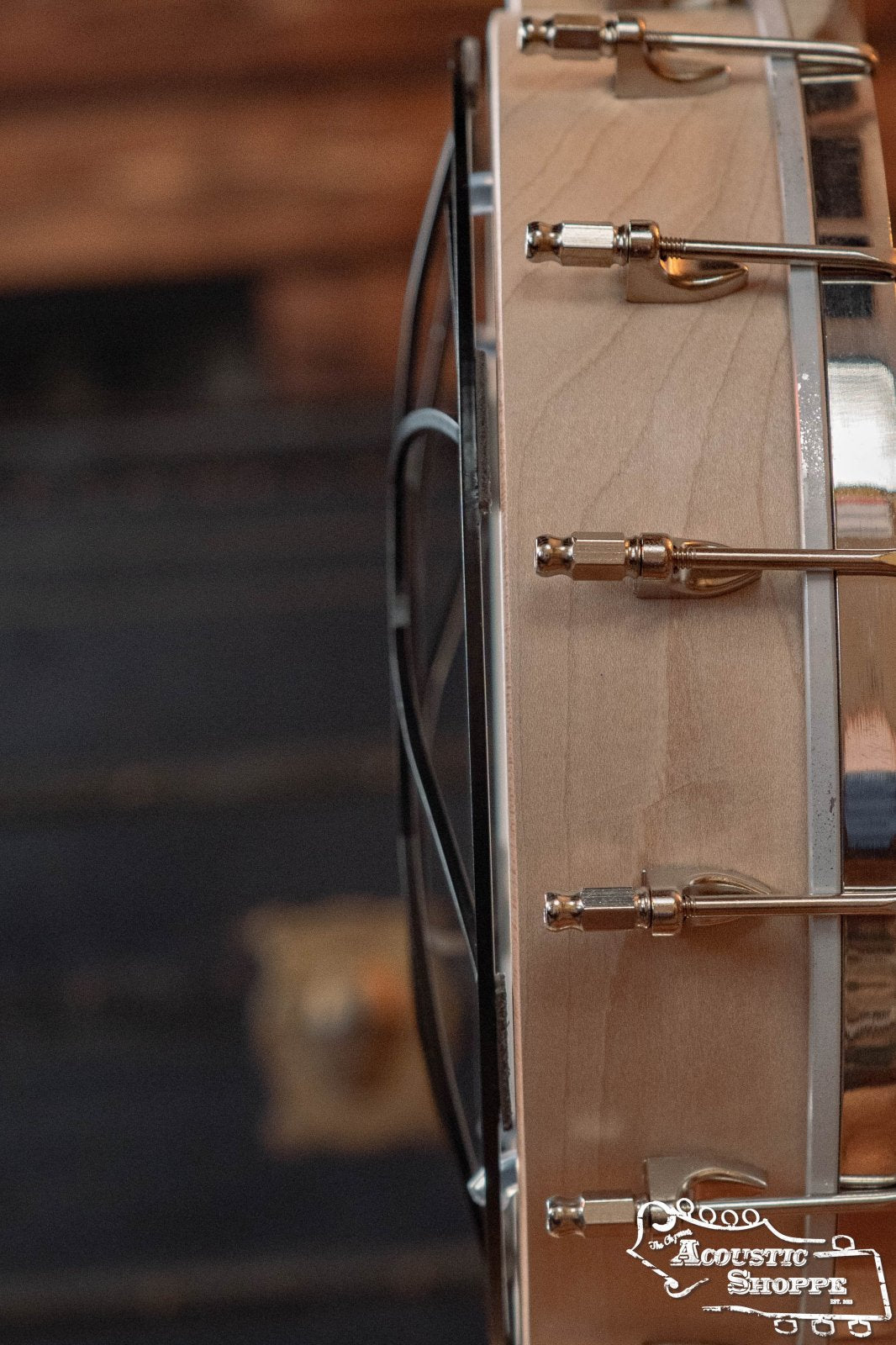 Close-up side view of a banjo, highlighting the wooden rim, metal brackets, and hardware—like how the Tone-Gard Open Back Banjo (11) by Tone-Gard improves tone and protection—with a blurred dark background and Acoustic Shoppe logo in the lower right.