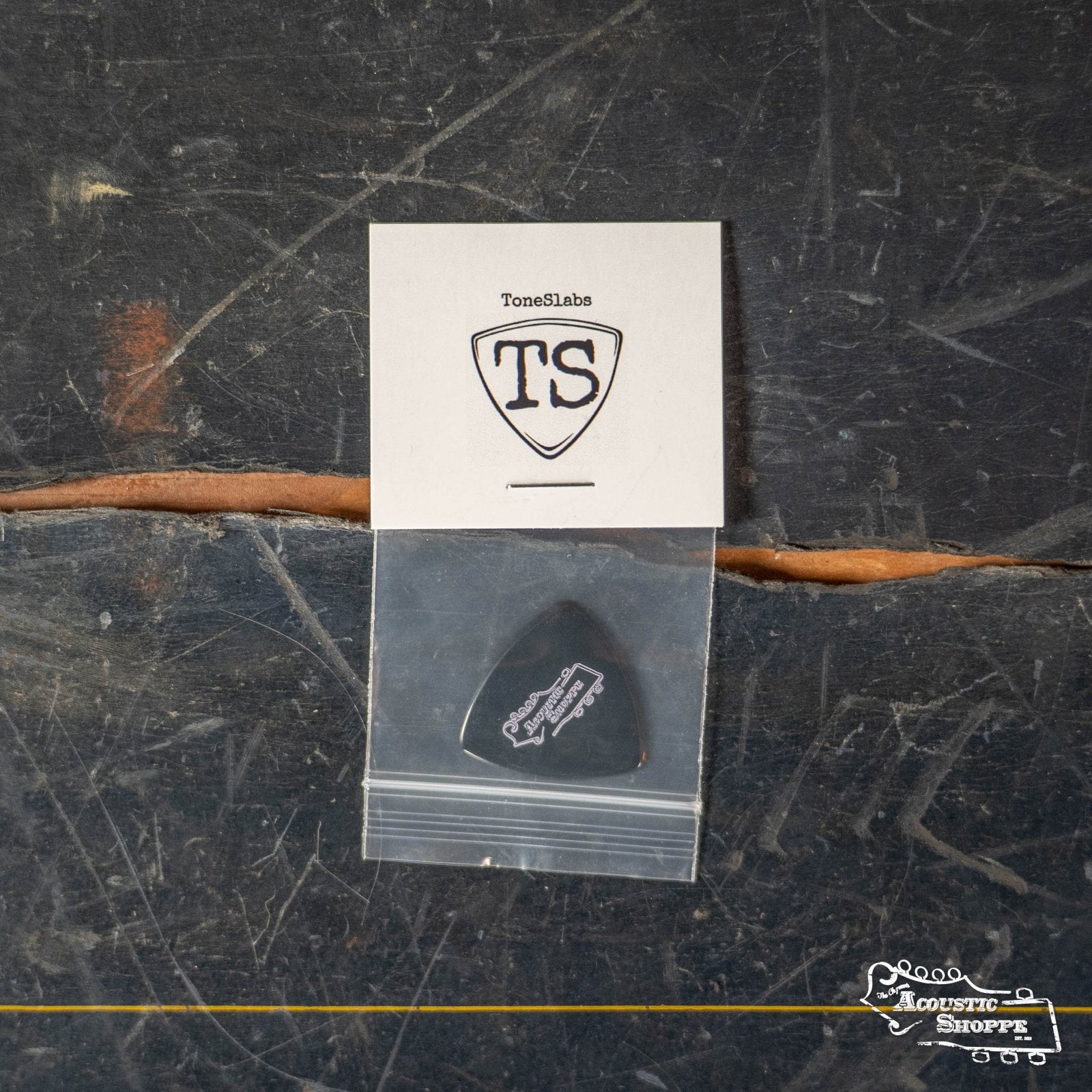 A ToneSlabs Darth Tone Series Tri Micro 1.5mm guitar pick in a small plastic bag sits on a scratched wooden surface, with a white card showing the ToneSlabs logo and TS, plus The Acoustic Shoppe logo in the bottom right corner.