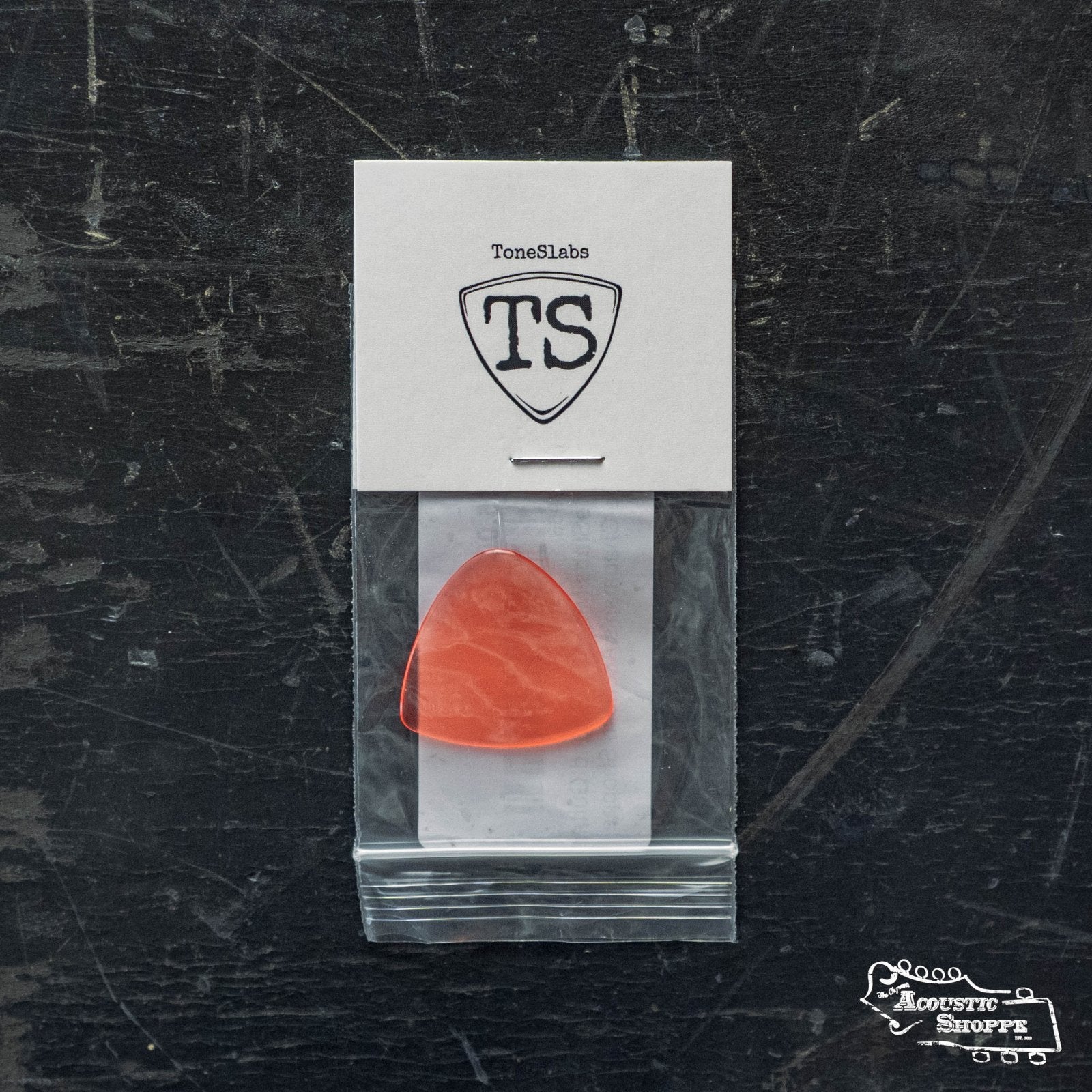 A ToneSlabs Mini Tri 1Round Guitar Pick (1.2mm) in orange comes in a clear bag attached to a branded card, displayed on a dark scratched surface with Acoustic Shoppe branding—ideal for acoustic guitar players.