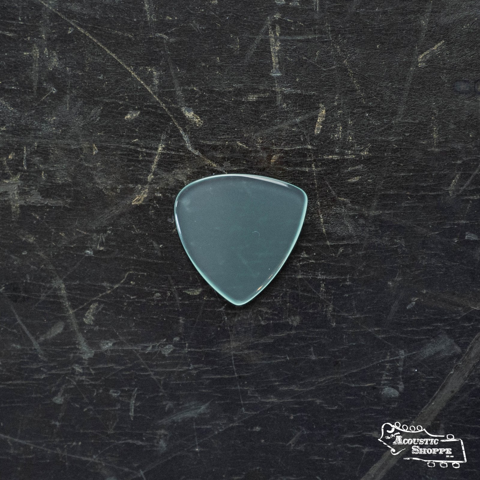 A translucent, light blue ToneSlabs TAS Custom Mini Tri 1Round Guitar Pick (1.3mm, Exclusive Color) rests on a dark, scratched surface with an Acoustic Shoppe logo—an essential guitar accessory from ToneSlabs.