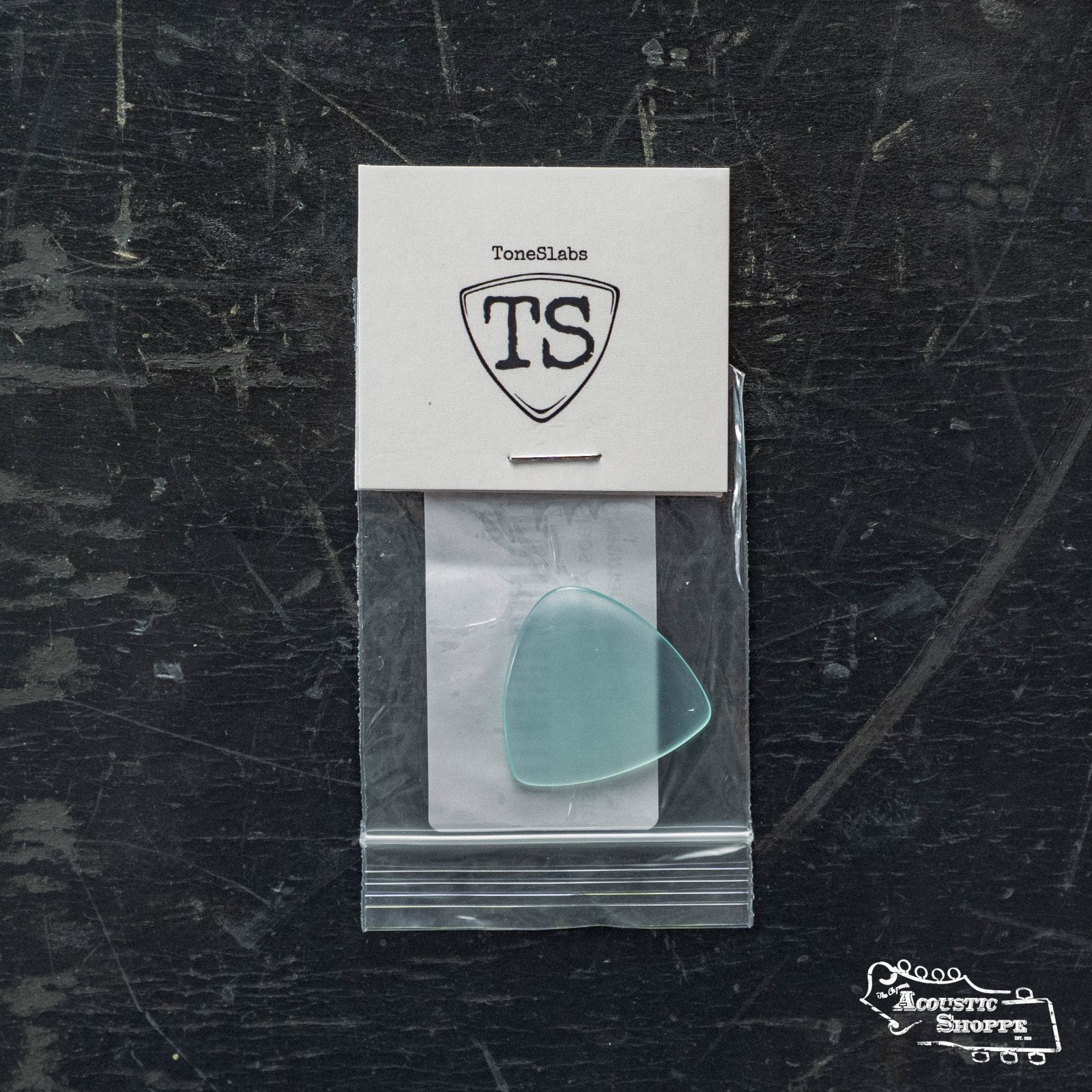 A ToneSlabs TAS Custom Mini Tri 1Round Guitar Pick - 1.3mm (Exclusive Color) is sealed in a bag and attached to a branded white card with a TS shield logo, resting on a dark surface featuring the Acoustic Shoppe logo.