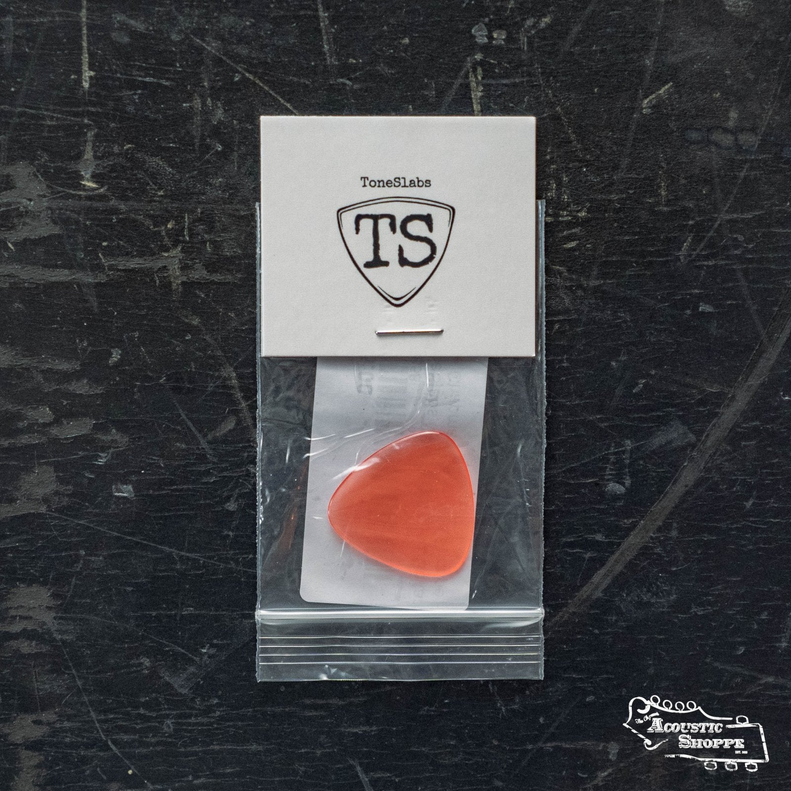 A red ToneSlabs Mini Tri 3Round Guitar Pick - 1.2mm comes in a small plastic bag, attached to a white ToneSlabs card with a TS pick outline, resting on a dark surface featuring the Acoustic Shoppe logo.