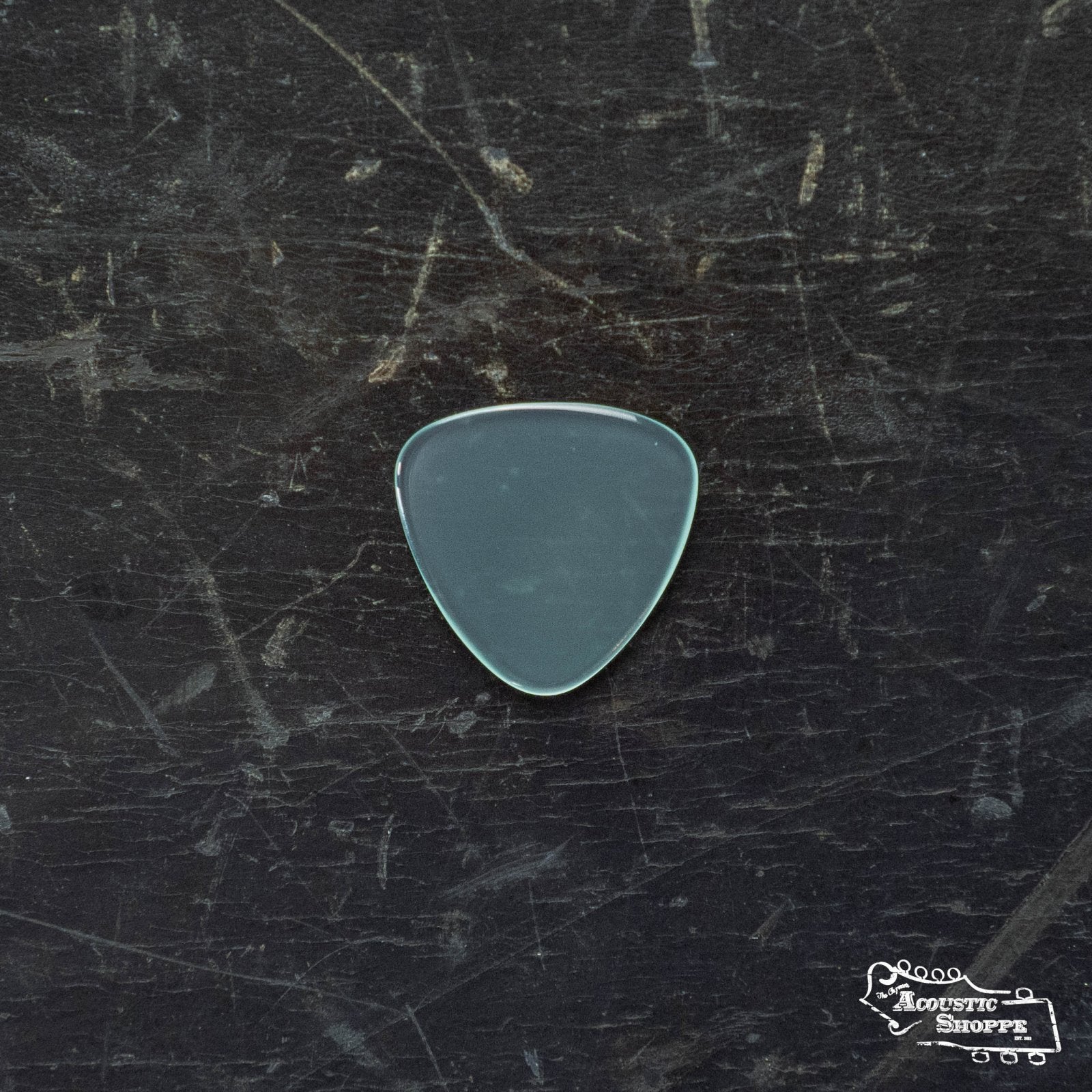 A translucent ToneSlabs TAS Custom Mini Tri 3Round Guitar Pick - 1.3mm (Exclusive Color) sits on a scratched, dark surface, with The Acoustic Shoppe logo in the bottom right, featuring essential guitar accessories.