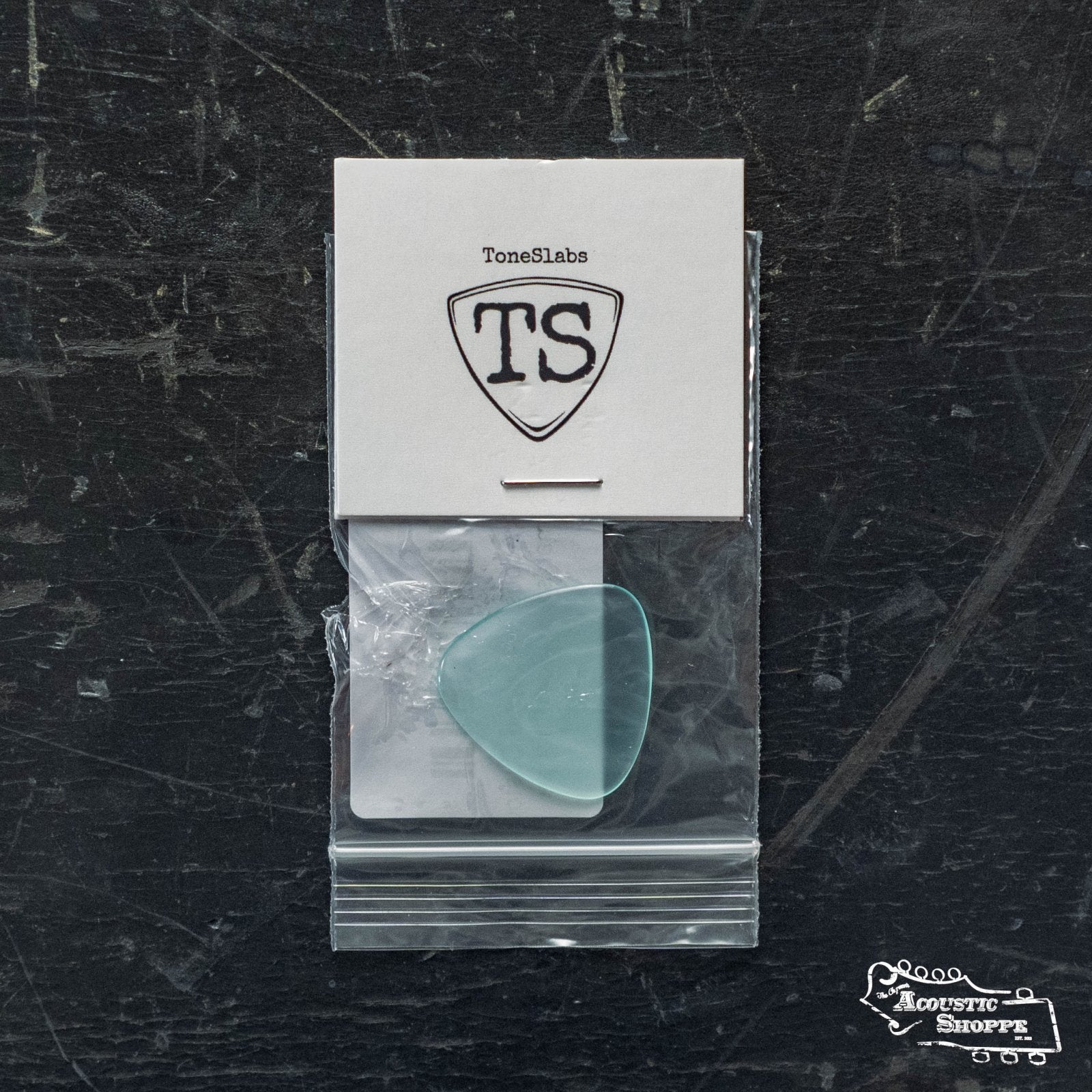 A translucent blue ToneSlabs TAS Custom Mini Tri 3Round 1.3mm guitar pick (exclusive color) by ToneSlabs sits in a clear bag on a dark, scratched surface with a TS logo card above and Acoustic Shoppe shown at the lower right—ideal for acoustic guitar accessories.