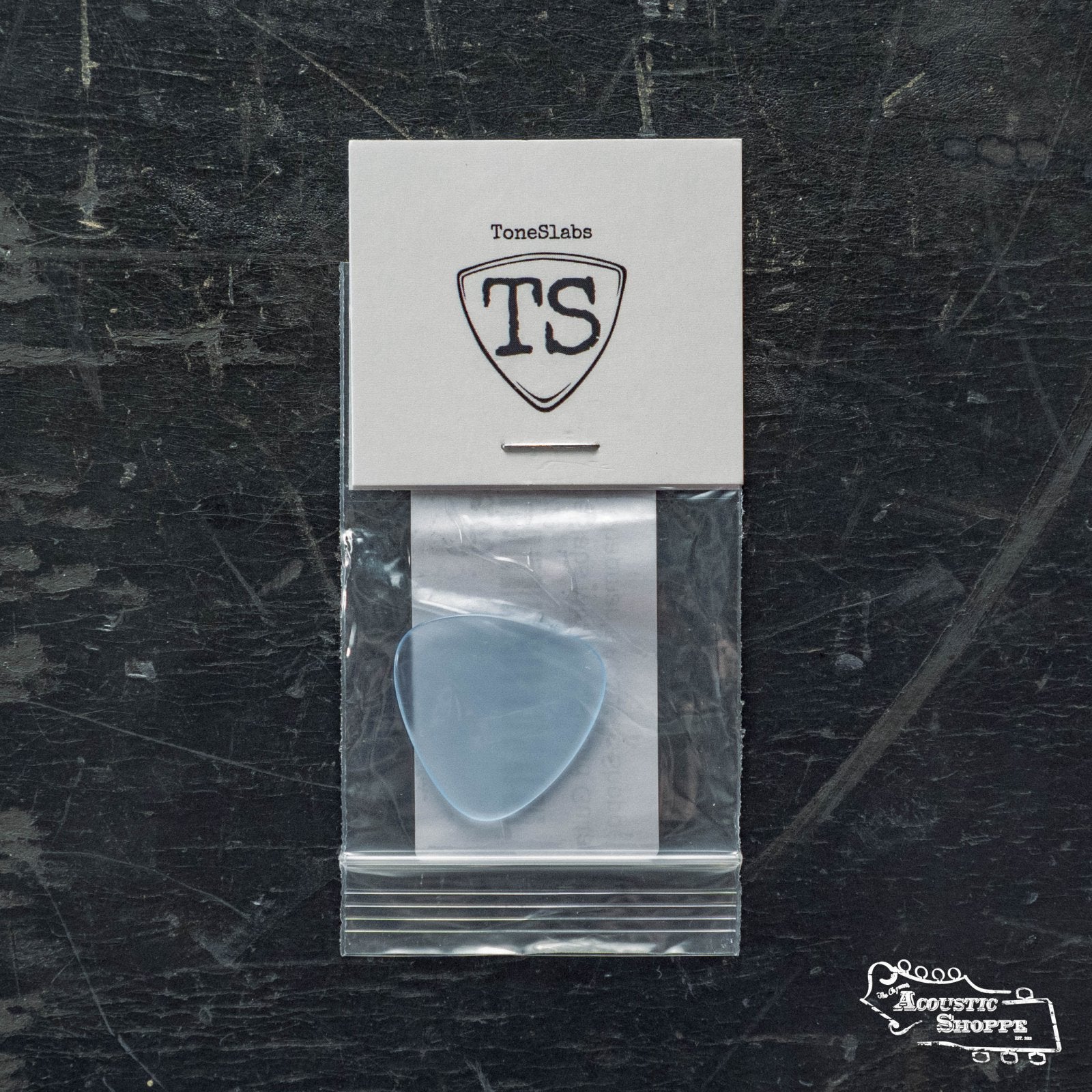 A translucent blue ToneSlabs Mini Tri 3Round Guitar Pick (1.4mm) is sealed in a small plastic bag, attached to a white ToneSlabs card with the TS logo. The Acoustic Shoppe logo appears in the lower right corner, set against a worn, dark surface.