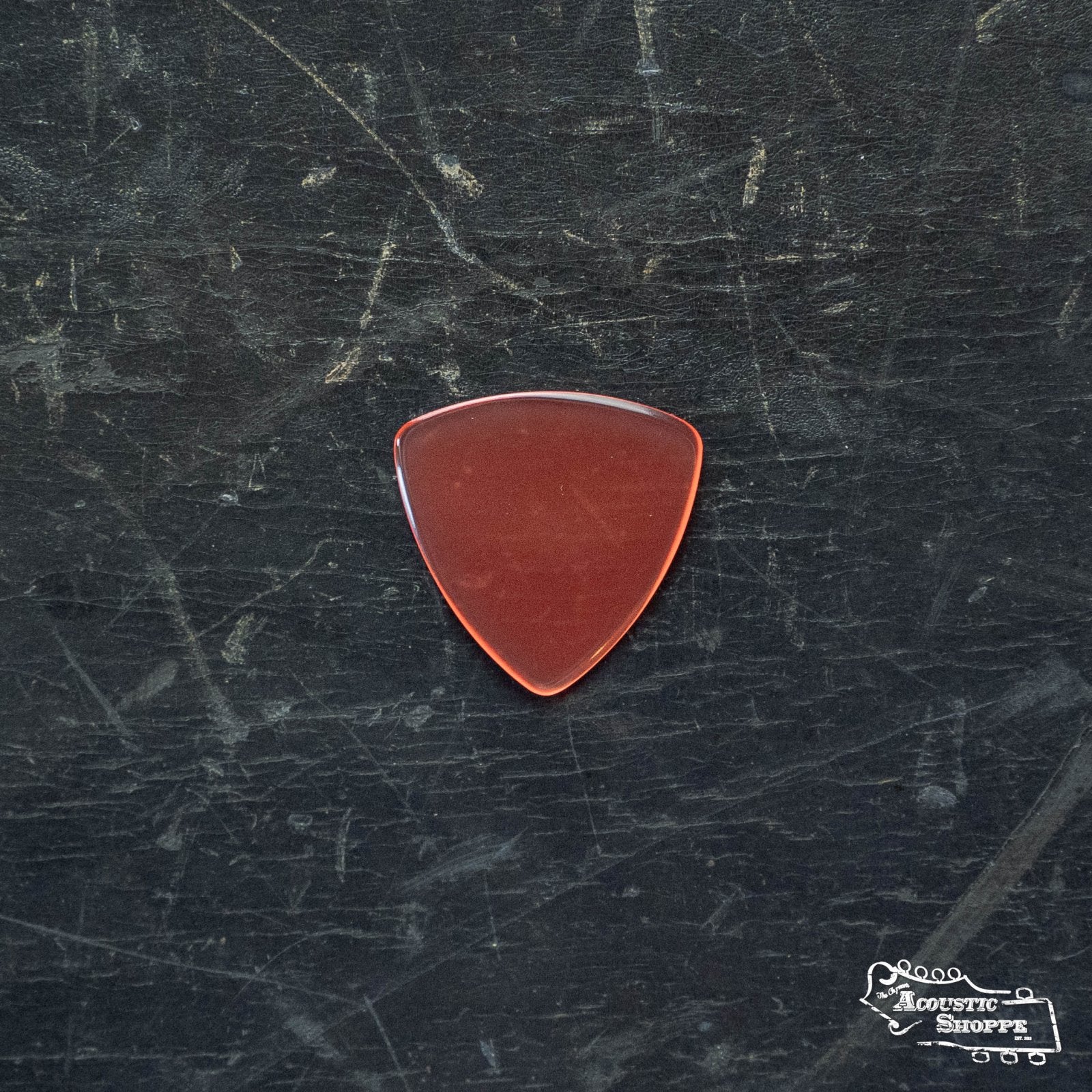A ToneSlabs Mini Tri Guitar Pick (1.2mm) in reddish-brown rests on a worn, dark, scratched surface with a small Acoustic Shoppe logo in the bottom right corner.