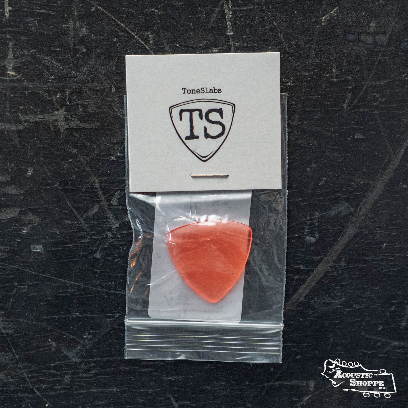 A ToneSlabs Mini Tri Guitar Pick - 1.2mm, in red, sits in a small plastic bag attached to a card with the TS logo on a scratched dark surface. The Acoustic Shoppe logo appears in the bottom right corner.