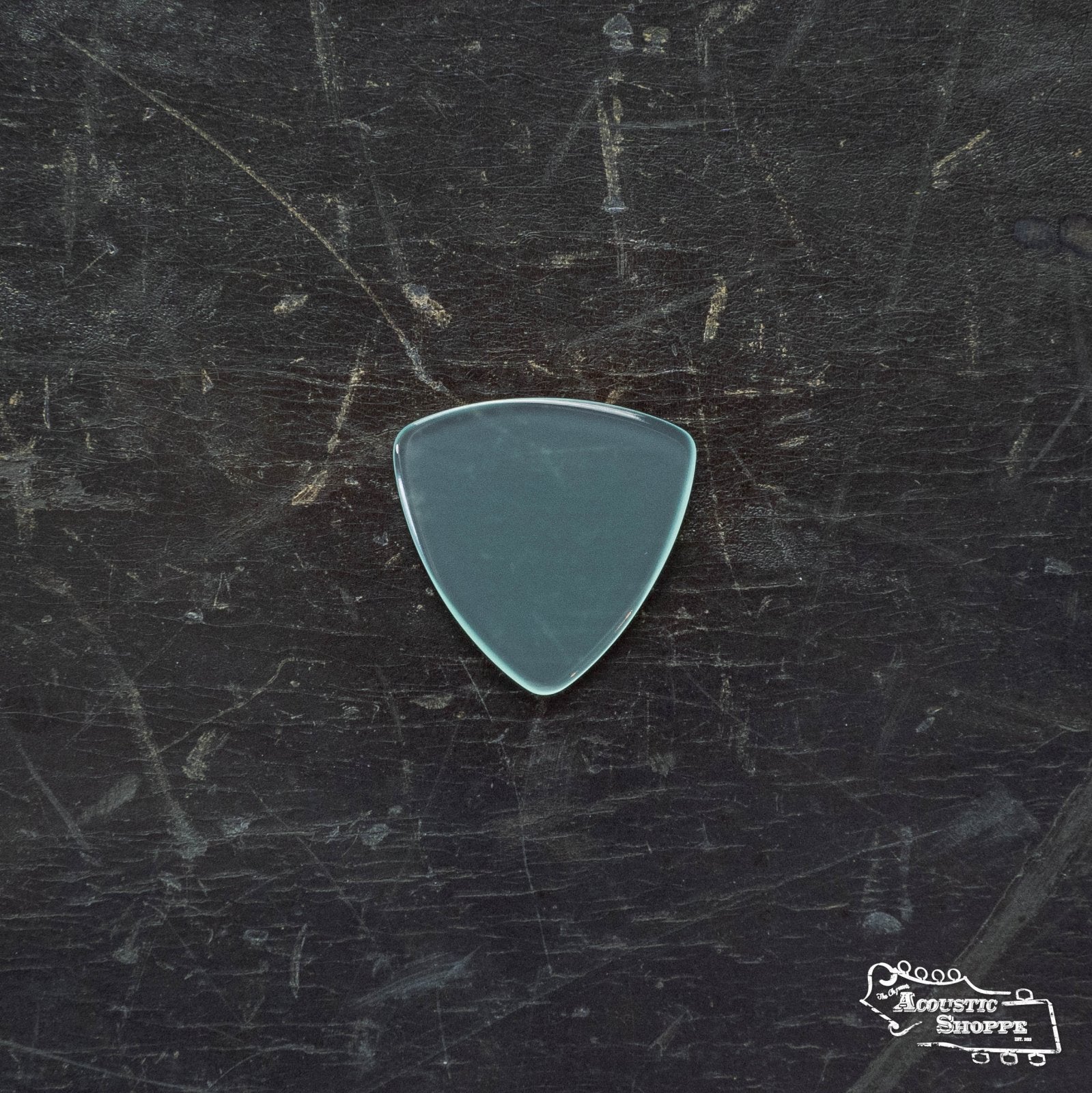 A translucent ToneSlabs Mini Tri Guitar Pick - 1.4mm with a triangular shape rests on a scratched, dark surface, featuring The Acoustic Shoppe logo in the bottom right corner.