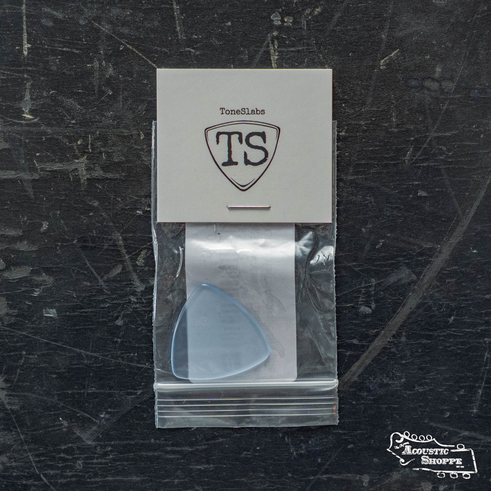 A ToneSlabs Mini Tri Guitar Pick - 1.4mm (blue) is shown in a clear bag with a ToneSlabs card marked TS, placed on a dark textured surface featuring the Acoustic Shoppe logo in the corner.