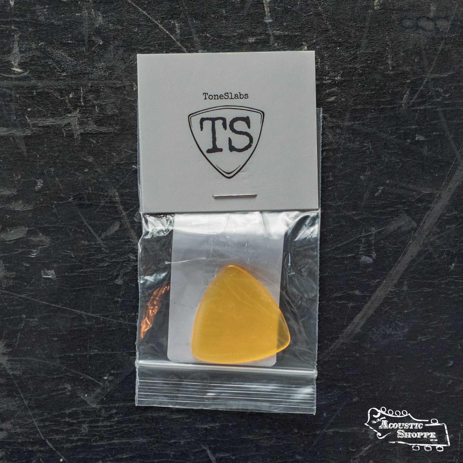 A 1.5mm ToneSlabs Mini Tri Guitar Pick in yellow, packaged with a TS logo card in a clear plastic bag, sits on a scratched dark surface with an Acoustic Shoppe logo. Ideal for acoustic players who prefer triangle picks.