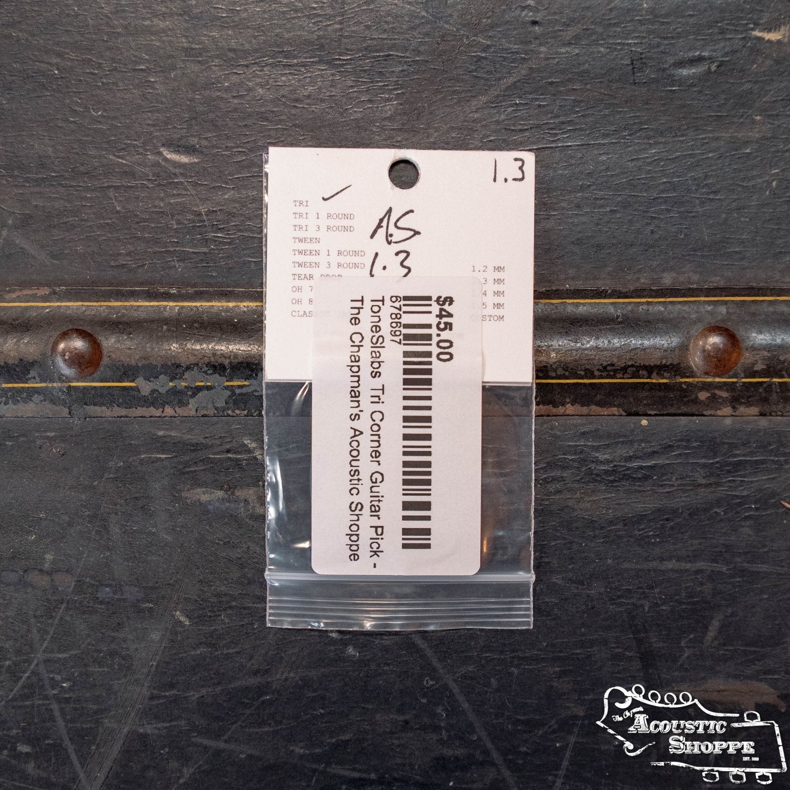 A ToneSlabs TAS Custom XL Tri Corner Guitar Pick, 1.3 mm, Exclusive Color/Signed, priced at $45 with barcode, is in a plastic bag labeled ToneSlabs and taped to a textured dark metallic surface at The Chapmans Acoustic Shoppe.