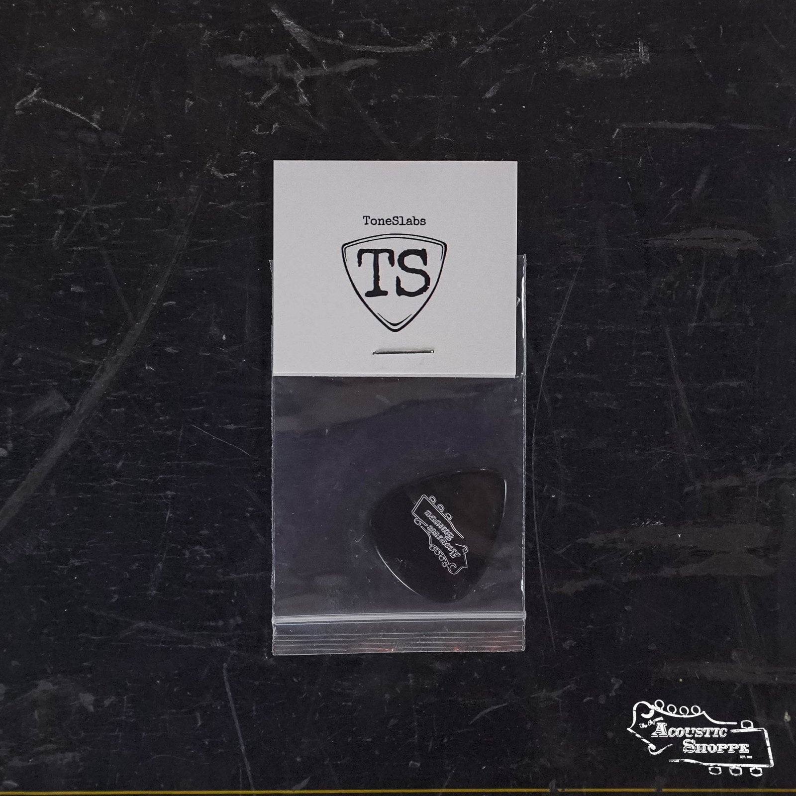The ToneSlabs Darth Tone Series Tear Drop Guitar Pick - 1.3mm comes in a small clear bag on a white card with the TS shield logo. The black pick features Acoustic Shoppe branding and is shown against a dark, textured background.