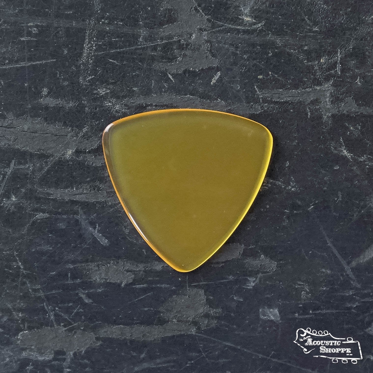 A translucent, amber ToneSlabs Frank Solivan Guitar Pick 1.5 with a sleek, rounded triangular shape rests on a dark, textured surface bearing faint scratches. The Acoustic Shoppe logo is subtly visible in the bottom right corner.