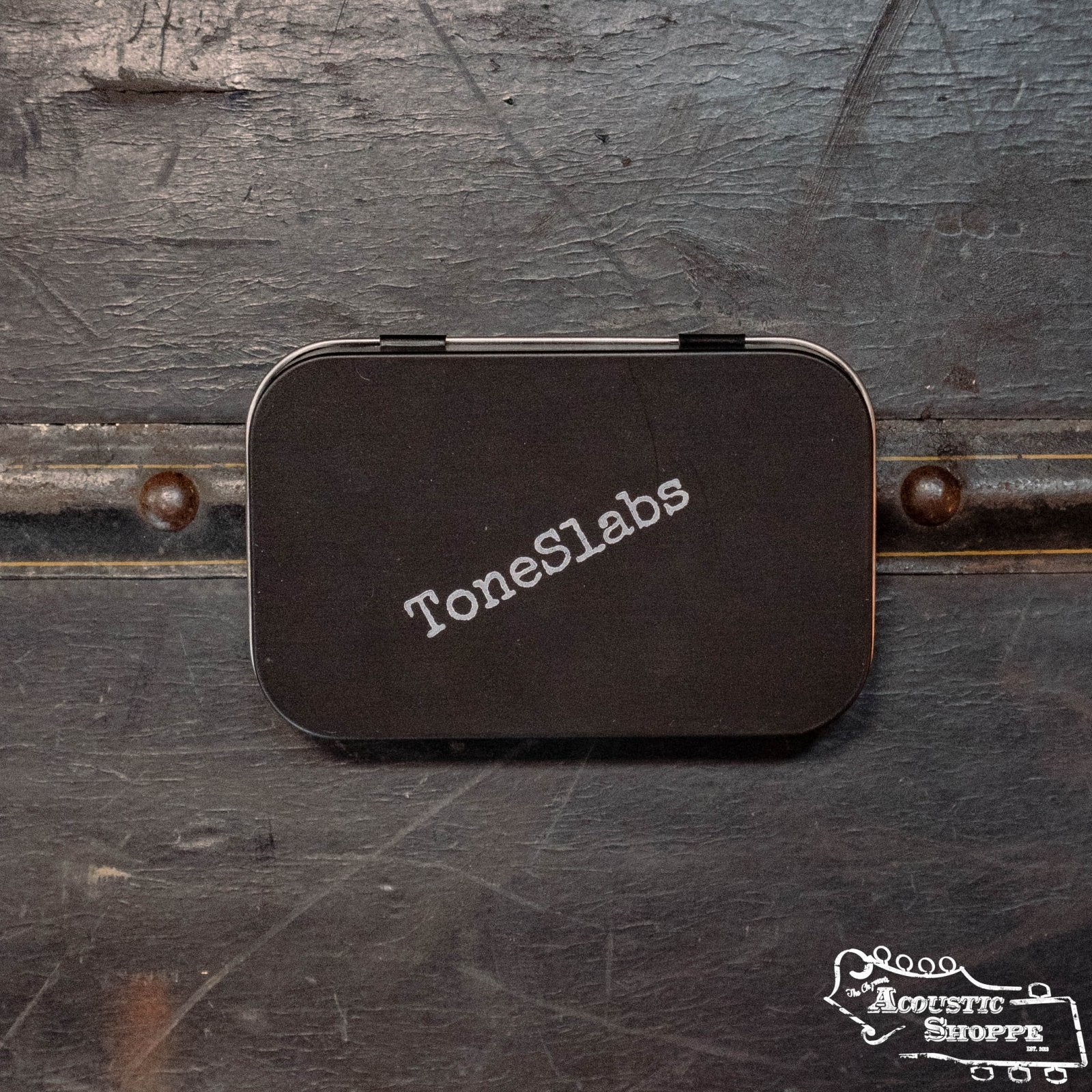 A small black rectangular ToneSlabs Pick Tin sits on a dark, worn surface. The Acoustic Shoppe logo, a boutique acoustic instrument retailer and Eastman dealer, appears in white at the lower right of the image.