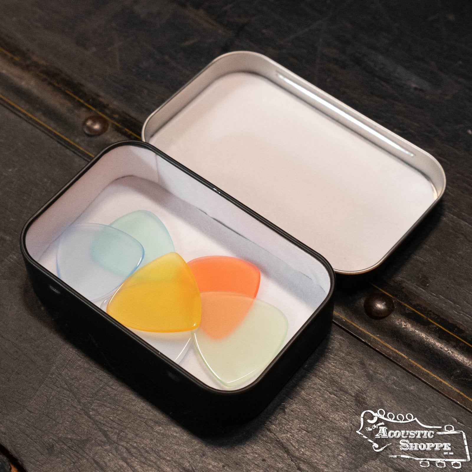 A ToneSlabs Pick Tin holds four translucent guitar picks—blue, yellow, orange, and green—on a dark textured surface. The Acoustic Shoppe, a premier music store and Eastman dealer, logo appears in the corner.