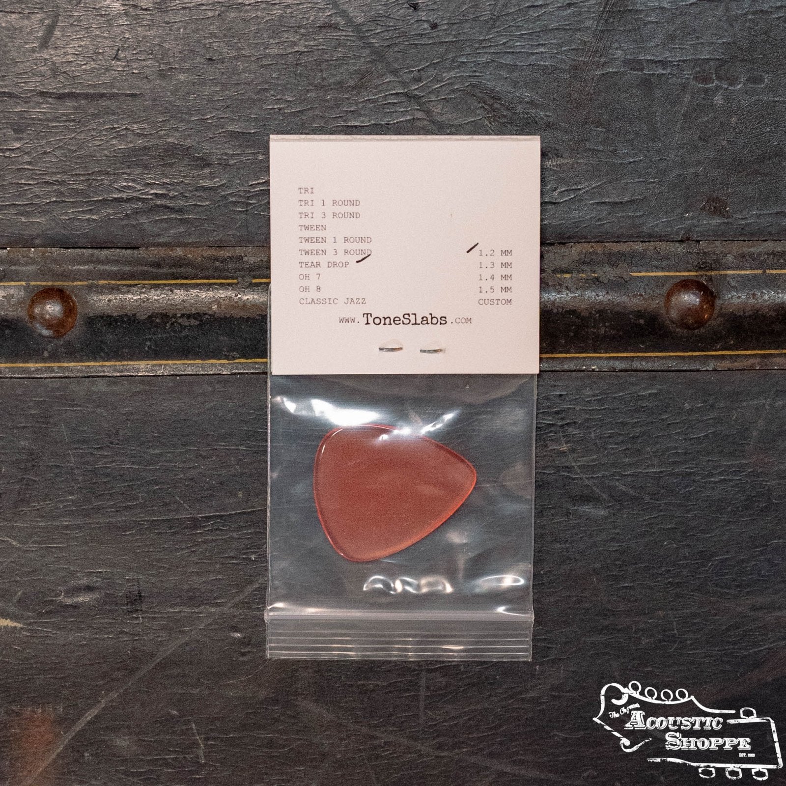 A reddish tortoise shell ToneSlabs Tear Drop Guitar Pick (1.2 mm) comes in a clear bag, attached to a branded white card with The Acoustic Shoppe logo, ideal for acoustic instruments and accessories, shown on a dark textured surface.