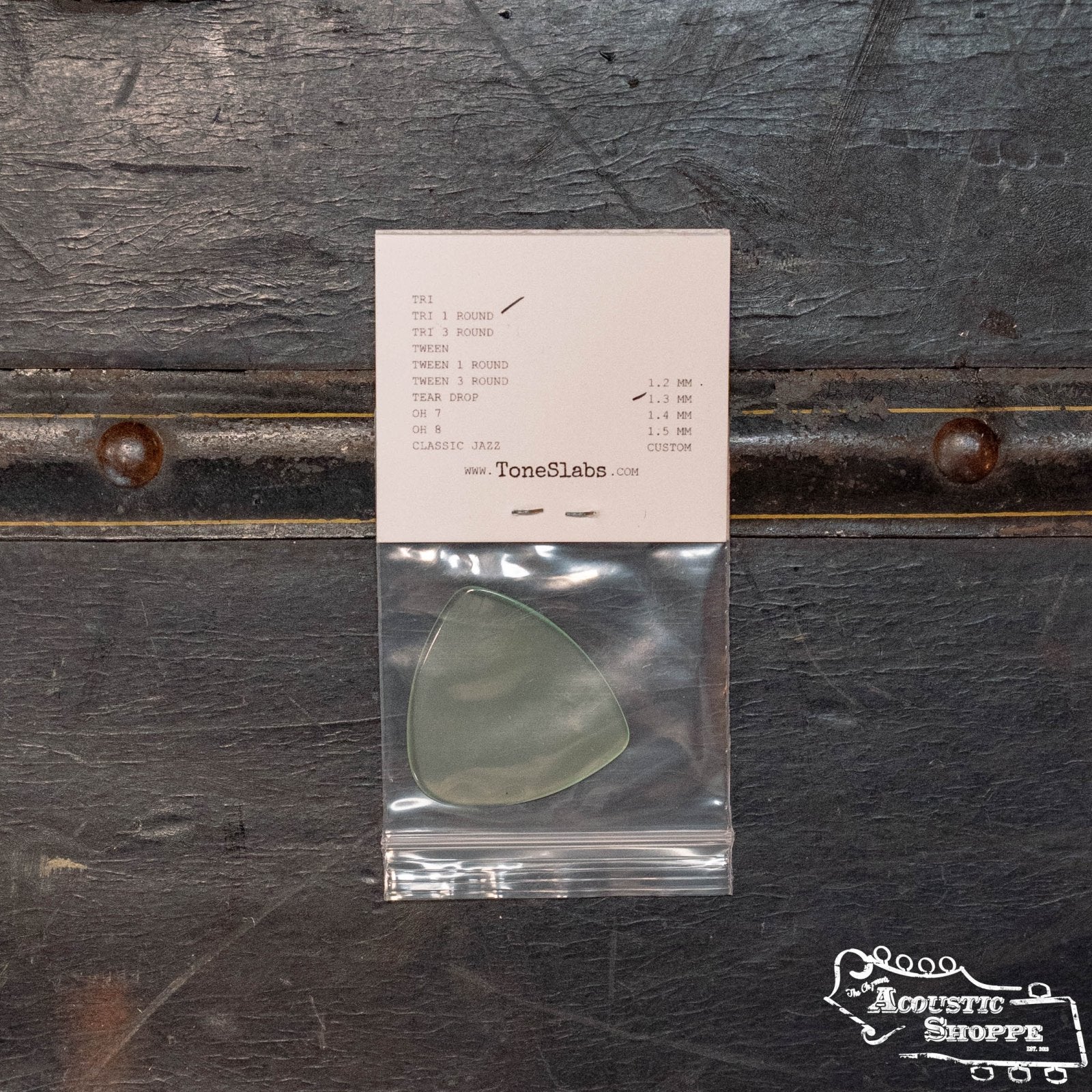 A ToneSlabs XL Tri Guitar Pick w/One Rounded Edge (1.3 mm) is shown in a small bag with an info card above it, resting on a dark textured surface featuring the Acoustic Shoppe logo, emphasizing ToneSlabs's superior tonal quality.