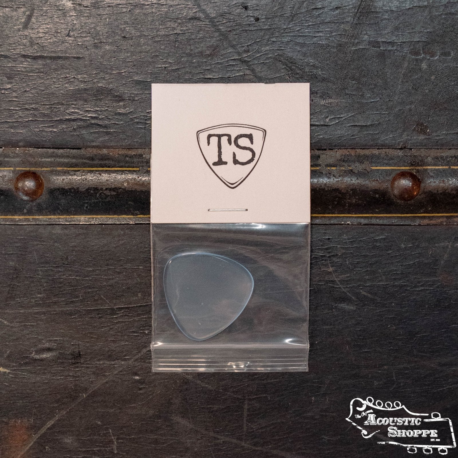 A ToneSlabs Tweener Triangle 3Rounded Guitar Pick - 1.4mm is in a clear plastic bag attached to a card with the “TS” logo, resting on a dark textured surface with metal accents. The Acoustic Shoppe logo is in the lower right corner.