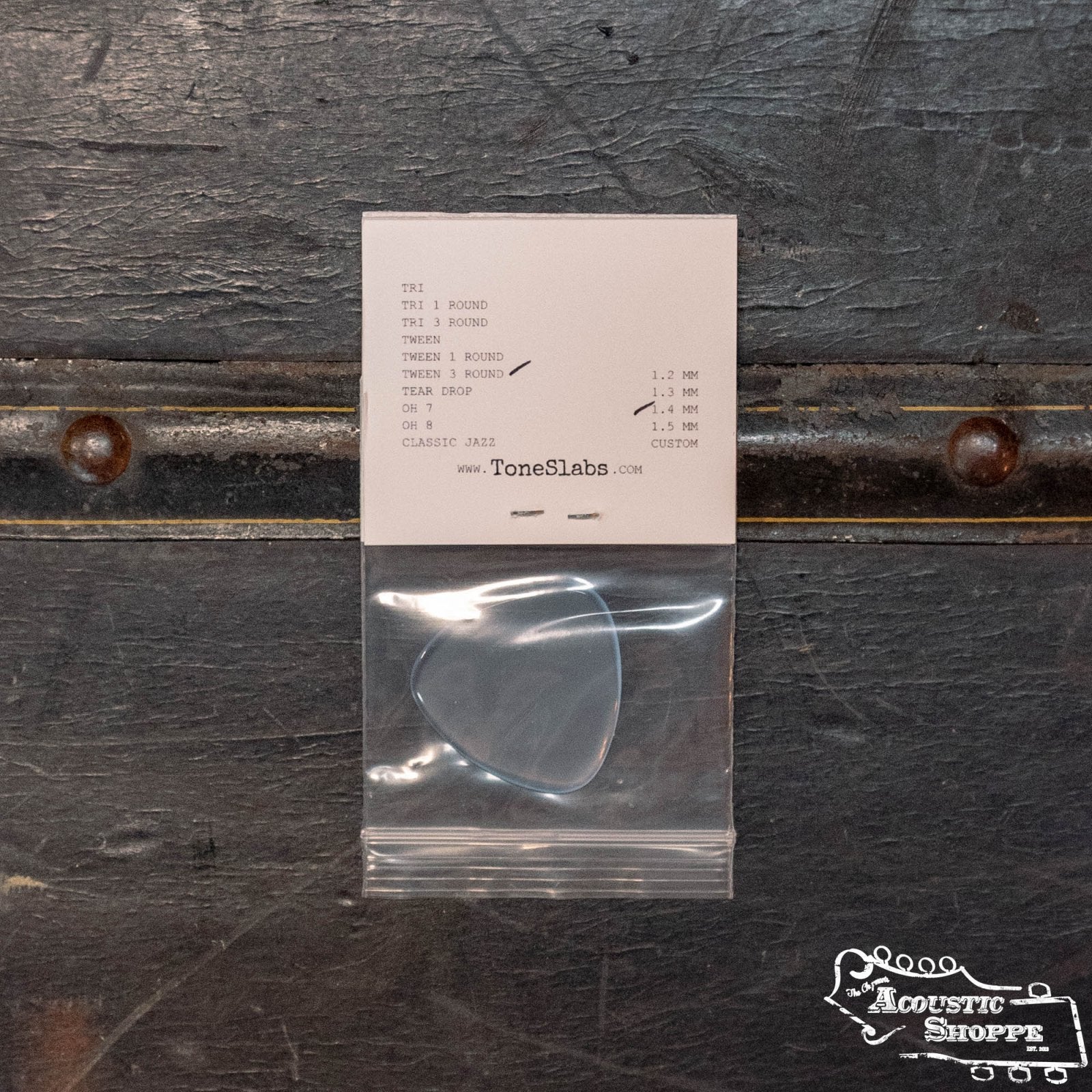 A ToneSlabs Tweener Triangle 3Rounded Guitar Pick - 1.4mm in a small plastic bag is attached to a card displaying model options and prices. The card, featuring the Acoustic Shoppe logo and ToneSlabs branding, sits on a dark textured surface.