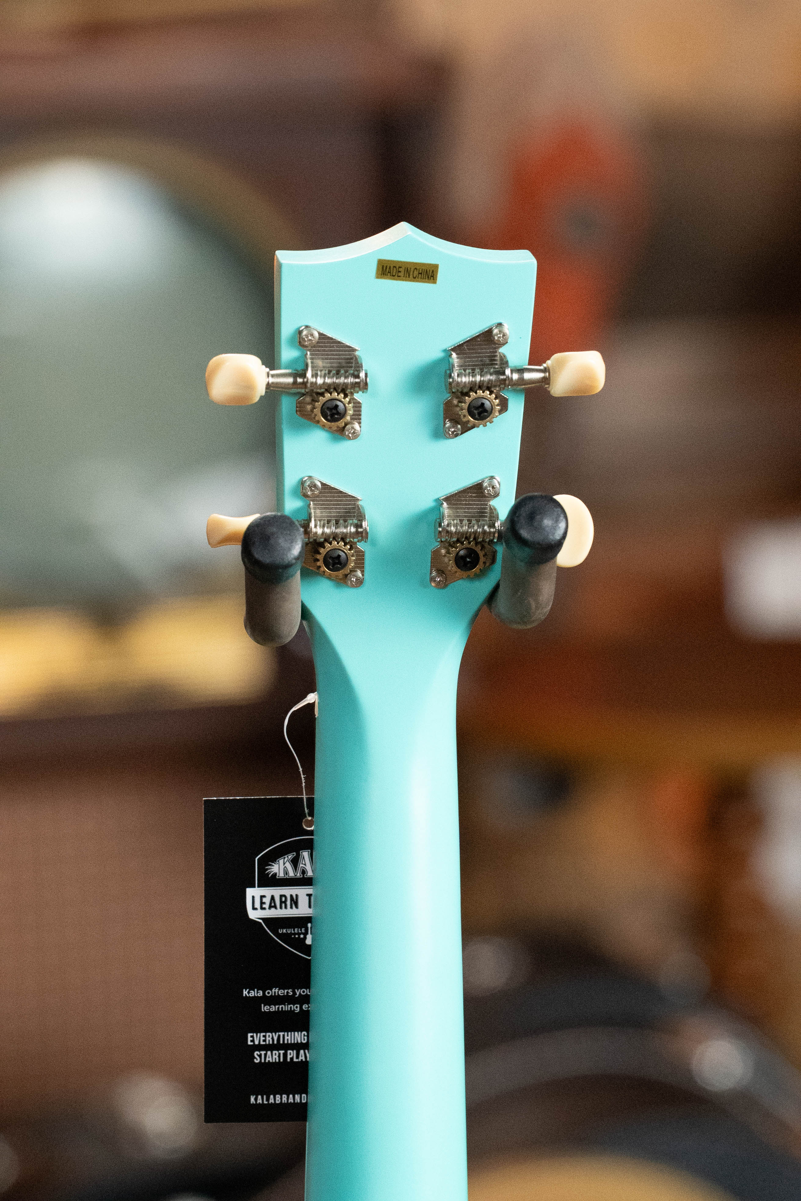 Aqua blue ukulele headstock with silver tuning pegs and cream-colored buttons against blurred background. - Ukadelic Seascape Soprano Ukulele - Kala Brand - Default Title
