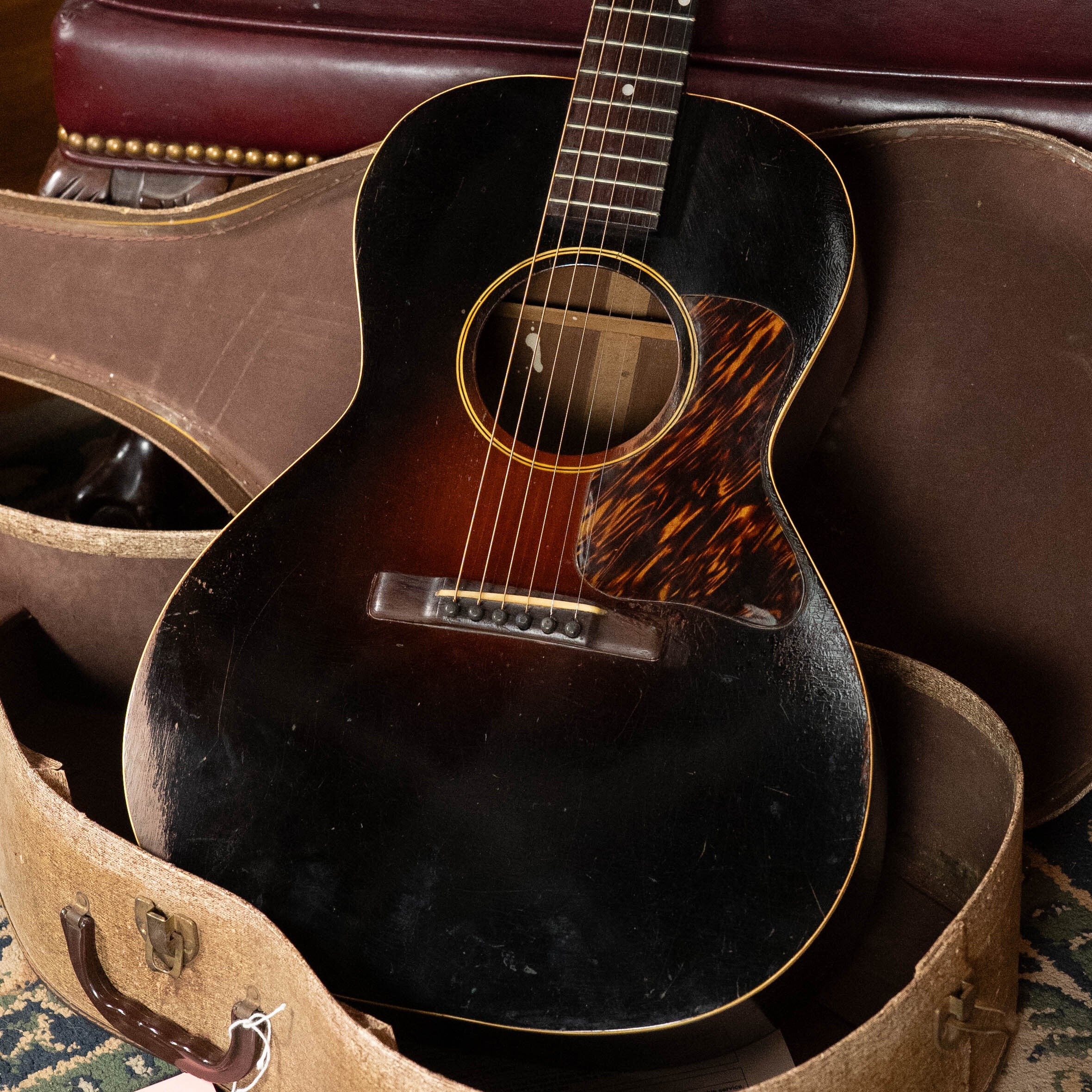 A (Used) 1933 Gibson LOO Adirondack/Mahogany OO Tobacco Sunburst Acoustic Guitar #550 by Gibson rests in an open, worn hardshell case on a carpeted floor.