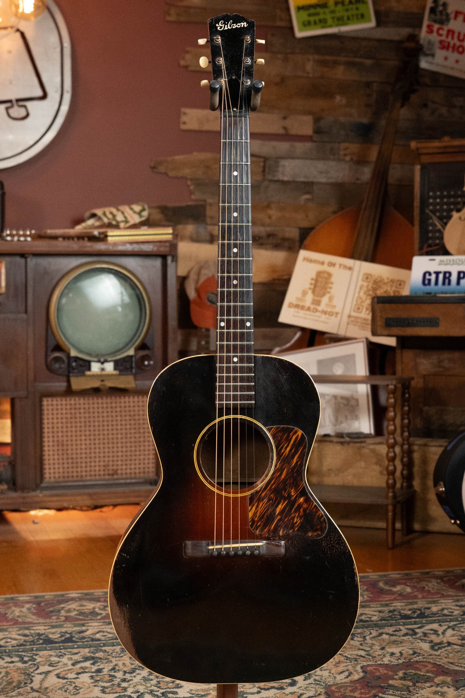 A (Used) 1933 Gibson LOO Adirondack/Mahogany OO Tobacco Sunburst Acoustic Guitar by Gibson stands upright in a cozy, retro room with wood-paneled walls, a patterned rug, and vintage musical gear in the background.
