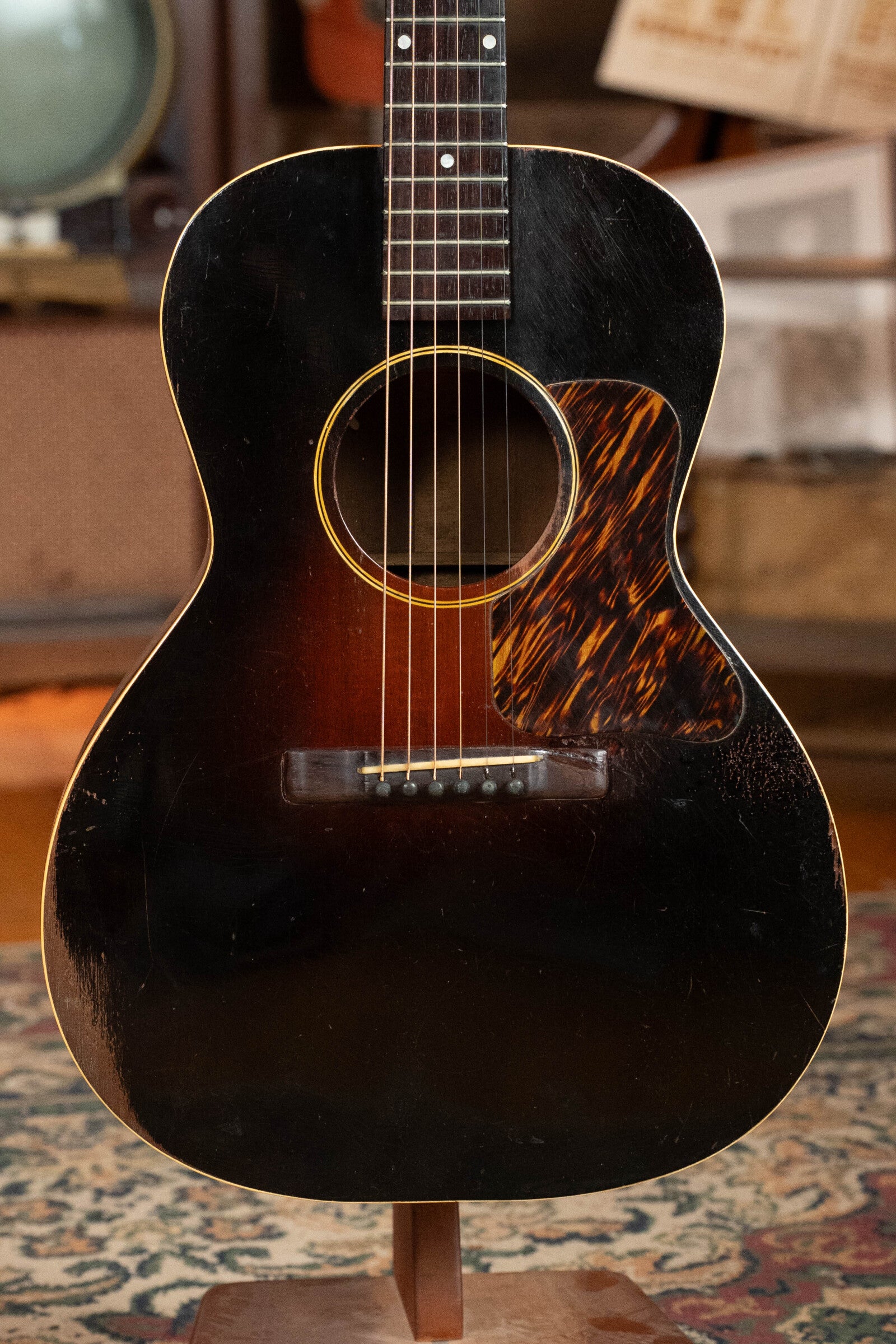 A (Used) 1933 Gibson LOO Adirondack/Mahogany OO Tobacco Sunburst Acoustic Guitar #550 by Gibson, with six strings and a tortoiseshell-style pickguard, is displayed upright on a stand in a cozy, warmly lit room.