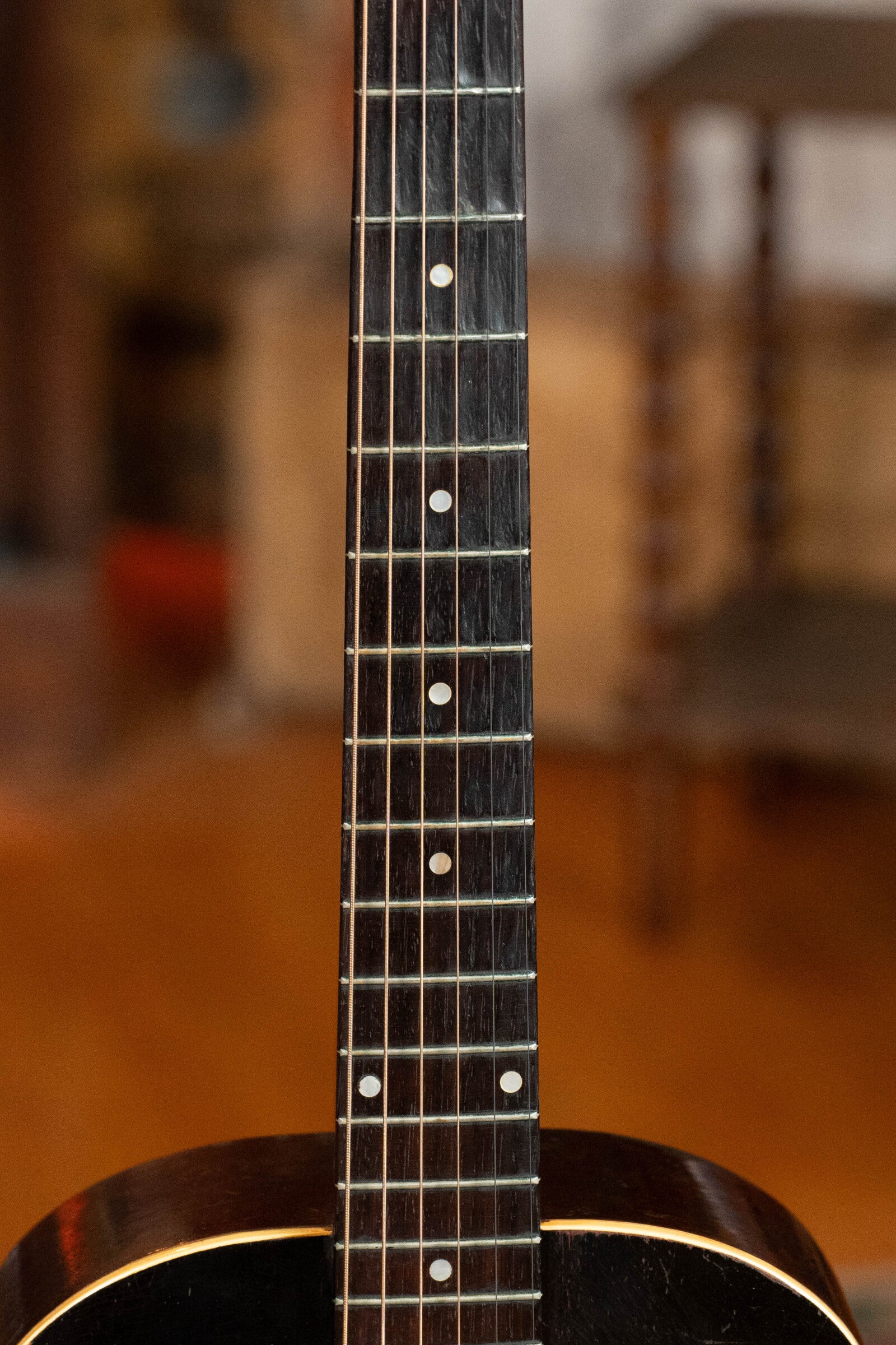 Close-up of the neck and upper body of a (Used) 1933 Gibson LOO Adirondack/Mahogany OO Tobacco Sunburst Acoustic Guitar #550 by Gibson, showing its dark fretboard with metal frets and white markers, set against a blurred indoor background.