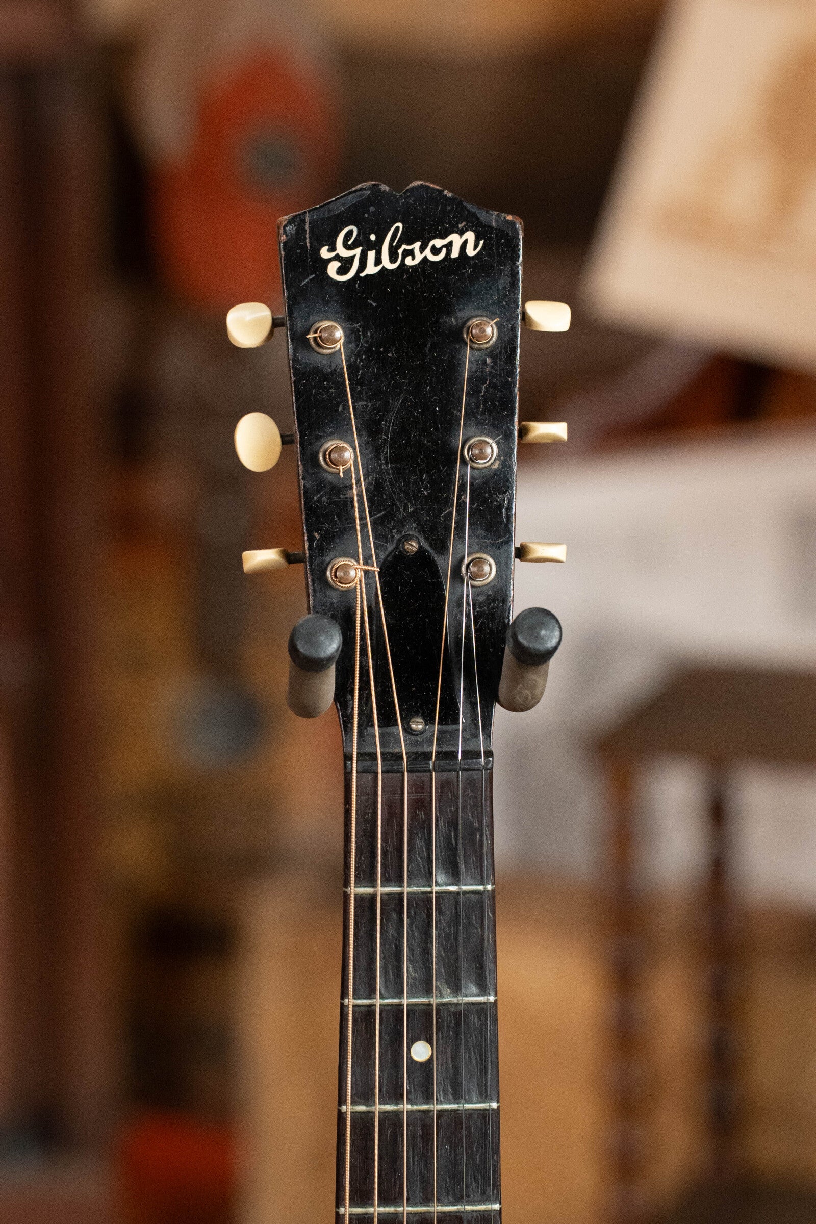 Close-up of the headstock on this used 1933 Gibson LOO Adirondack/Mahogany OO Tobacco Sunburst acoustic guitar, featuring tuning pegs and fretboard, with a blurred room and assorted objects in the background.