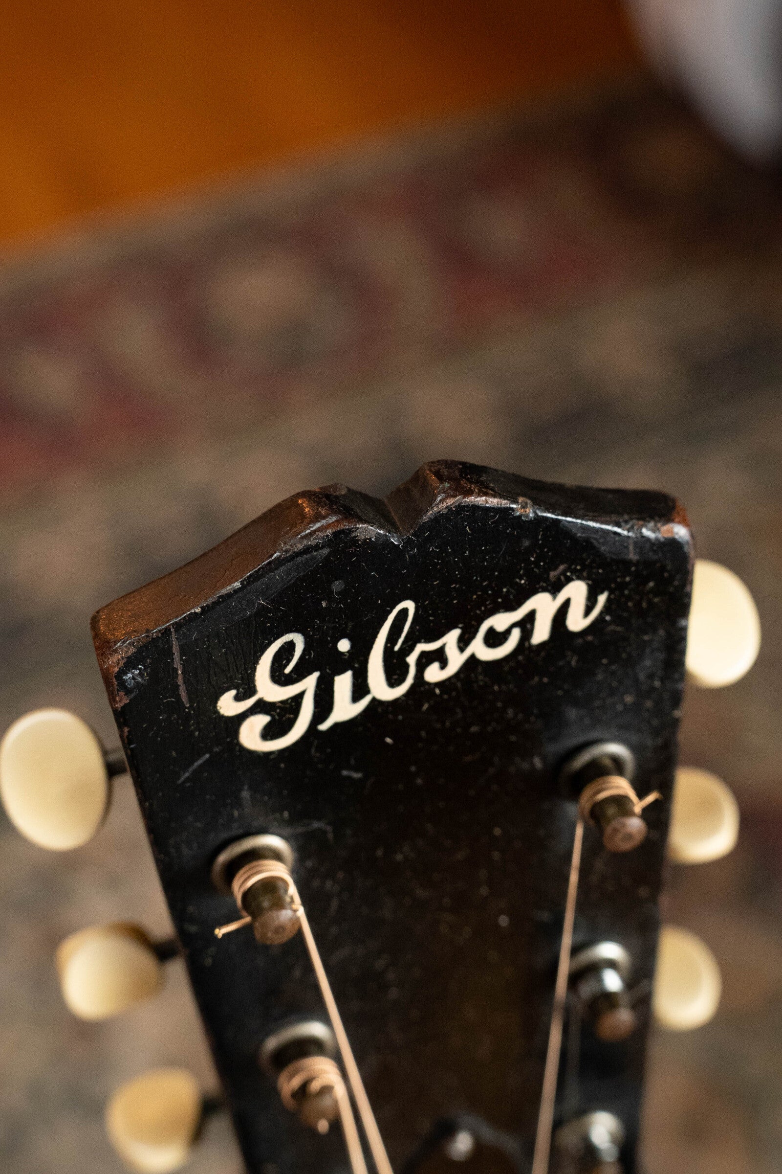 Close-up of the headstock of a black 1933 Gibson LOO Adirondack/Mahogany OO Tobacco Sunburst Acoustic Guitar #550 with cream tuning pegs and white Gibson logo, set against a blurred patterned background.