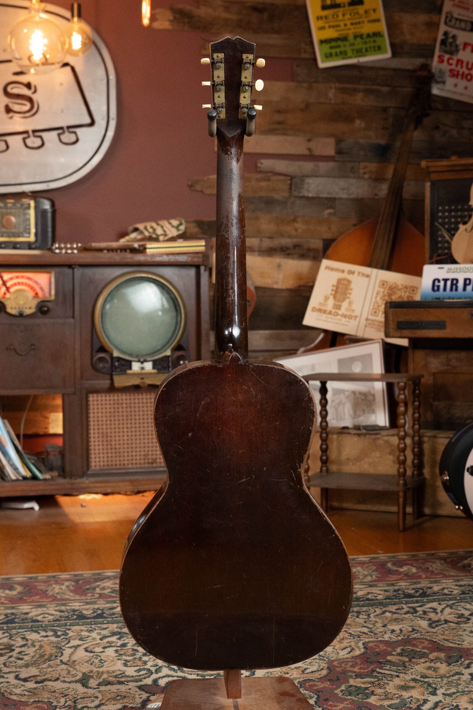A (Used) 1933 Gibson LOO Adirondack/Mahogany OO Tobacco Sunburst Acoustic Guitar #550 by Gibson, shown from the back on a patterned rug in a cozy retro-themed room with wooden walls, a TV, and other musical instruments in the background.