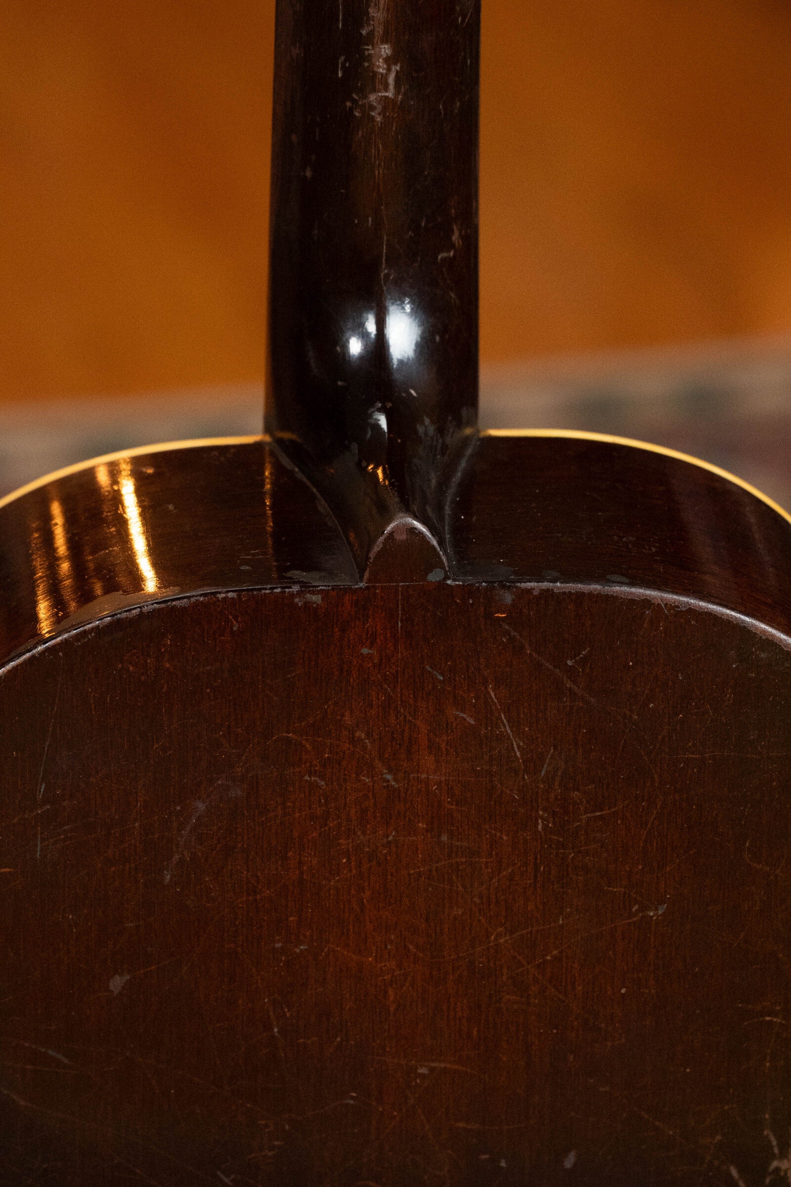 Close-up of the neck joint on a (Used) 1933 Gibson LOO Adirondack/Mahogany OO Tobacco Sunburst Acoustic Guitar #550, showing its glossy reddish-brown finish and vintage wear against a blurred orange-brown background.