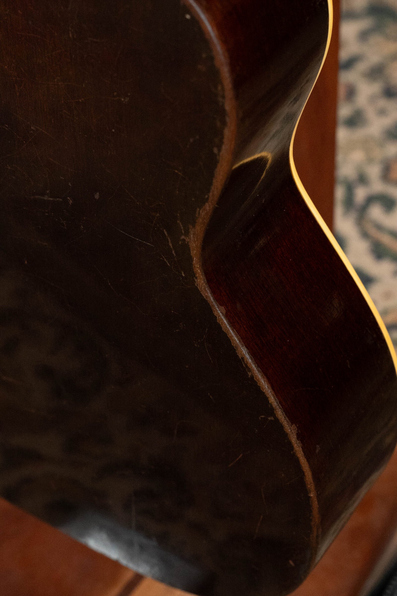 Close-up of the side and back of a (Used) 1933 Gibson LOO Adirondack/Mahogany OO Tobacco Sunburst Acoustic Guitar #550 by Gibson, showing scratches and worn edges that emphasize its vintage condition. Part of a patterned rug appears in the background.